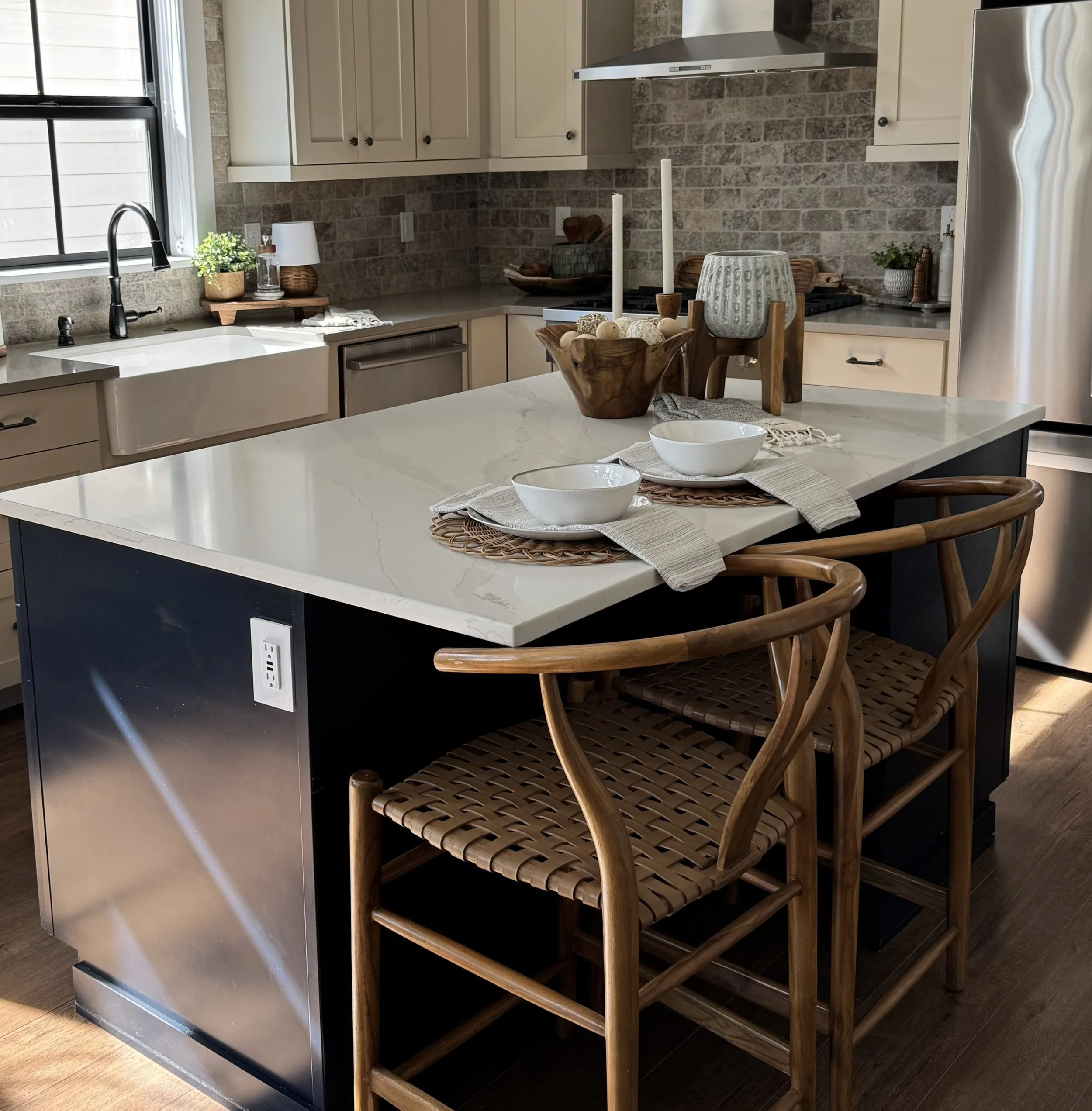 A modern kitchen with beige cabinetry, gray brick backsplash, and stainless steel appliances. A kitchen island has two white bowls on textured placemats, and wooden chairs with woven seats are positioned around it. Decorations include potted plants and candles.