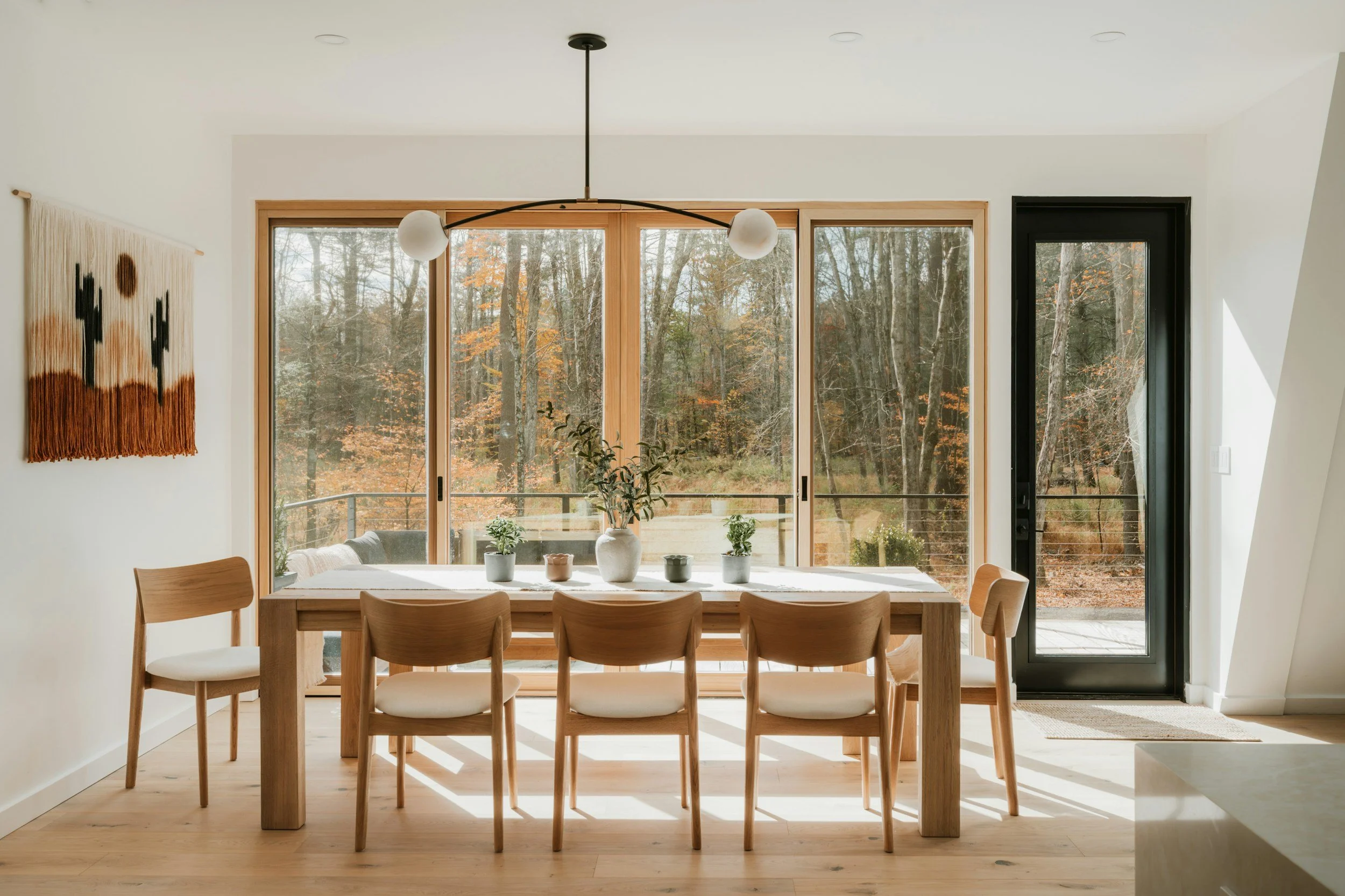 Interior dining room with large windows revealing trees with fall foliage, wooden dining table with six chairs, decorative plants, and a modern pendant light fixture.