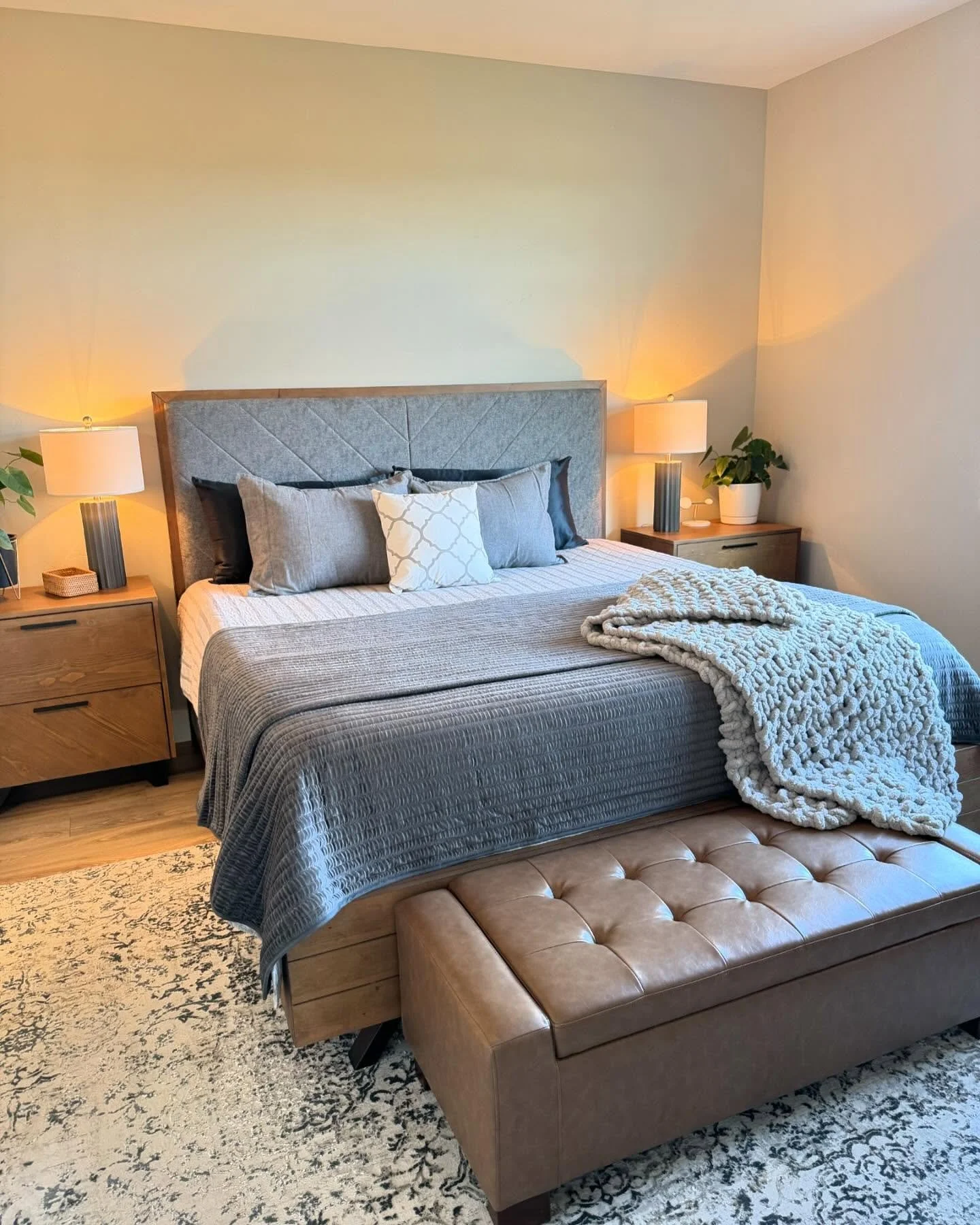 A cozy bedroom with a large bed, gray quilted bedspread, multiple pillows, and a chunky white blanket. Two nightstands with lamps and plants flank the bed. A tan leather tufted bench is at the foot of the bed, and a patterned rug covers the wooden floor.