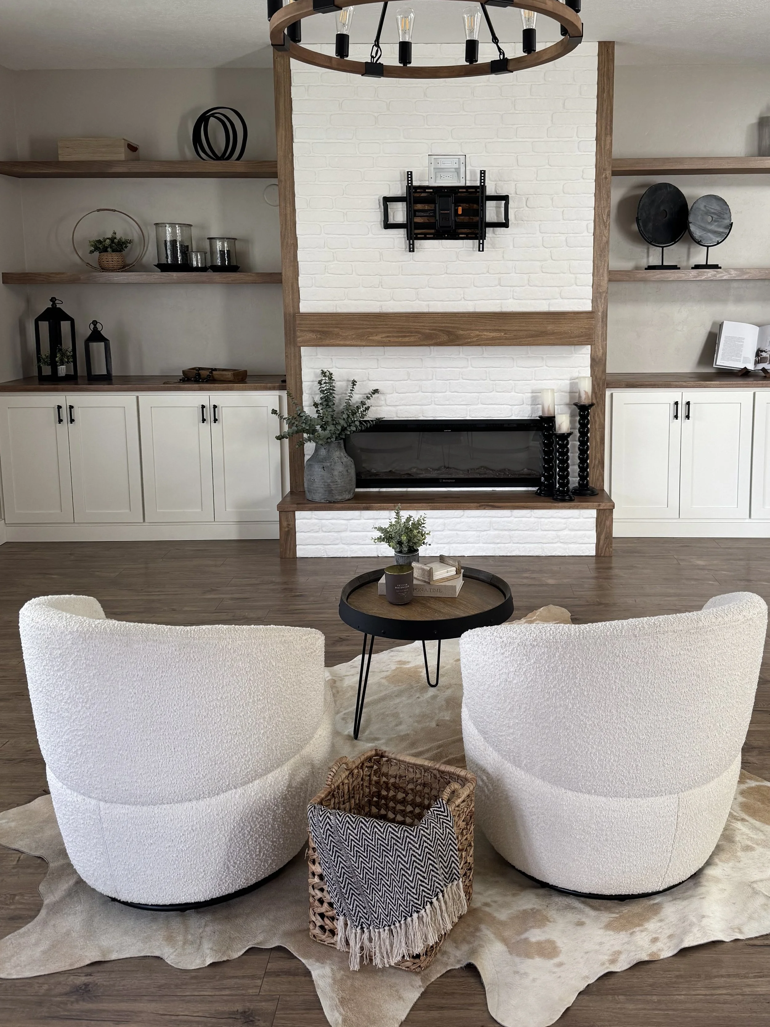 Living room with two white, textured armchairs around a small round black coffee table on a beige cowhide rug. Behind them is a white brick fireplace with a wooden mantle, decorated with candles, a vase with greenery, and black candle holders. To the left and right of the fireplace are built-in wooden shelves with decorative items, including vases, candles, books, and art pieces. There is a light wood floor and neutral wall colors.