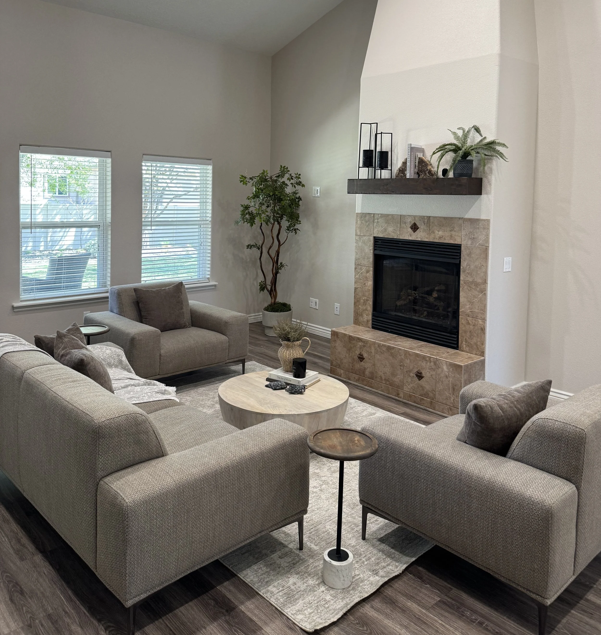 Living room with beige L-shaped sofa, round wooden coffee table, gray armchair, potted plant, fireplace with tile surround, decorative items, and windows with blinds.