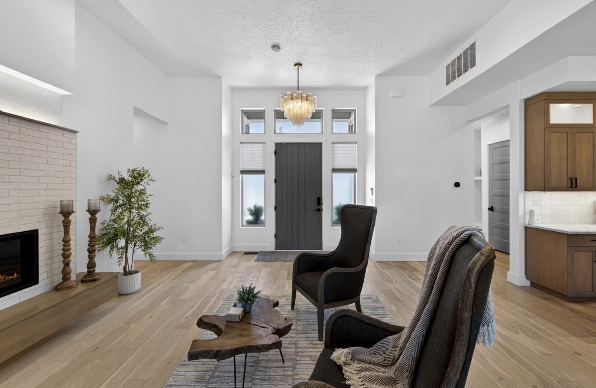 Living room with wooden flooring, a black front door, tall windows, a chandelier, a fireplace, a potted plant, and two armchairs with a small wooden table in the middle.