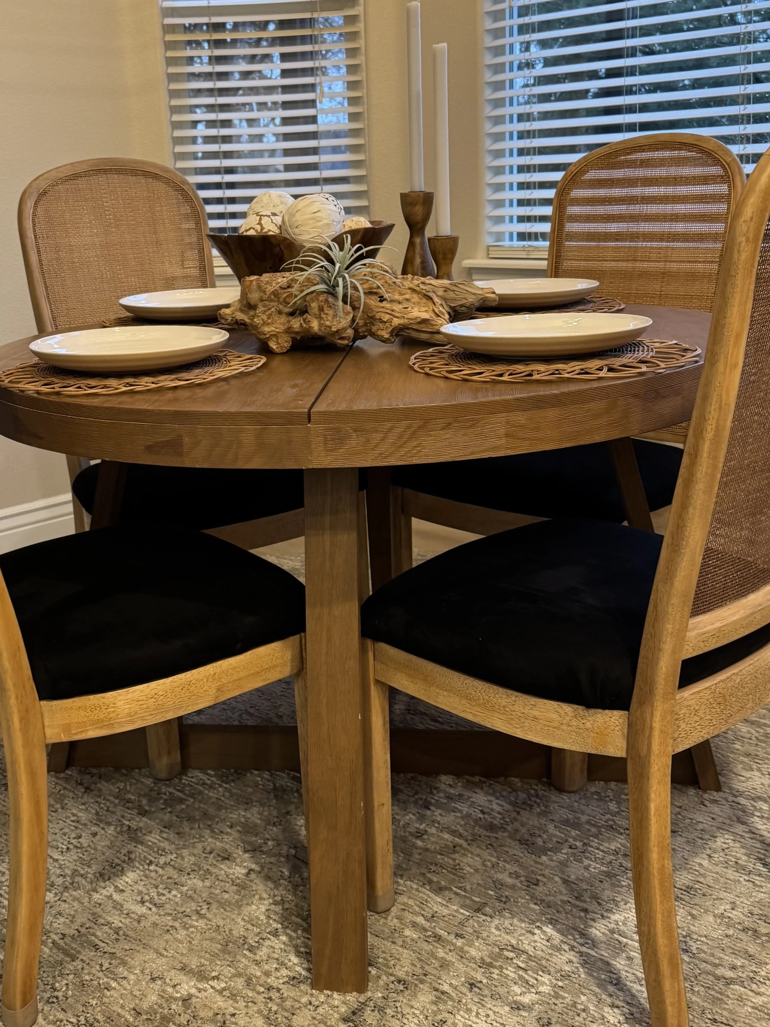 A round wooden dining table set with four white plates and brown woven placemats, decorated with a centerpiece of driftwood, a small air plant, a bowl with decorative balls, and two tall candle holders with white candles. The table is surrounded by wooden and woven chairs, with window blinds and trees visible in the background.
