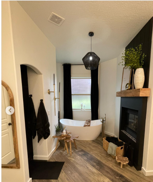Modern bathroom with a freestanding bathtub, a black fireplace with a wooden mantel, a large mirror, and a window with black curtains, decorated with a plant and minimalist accessories.
