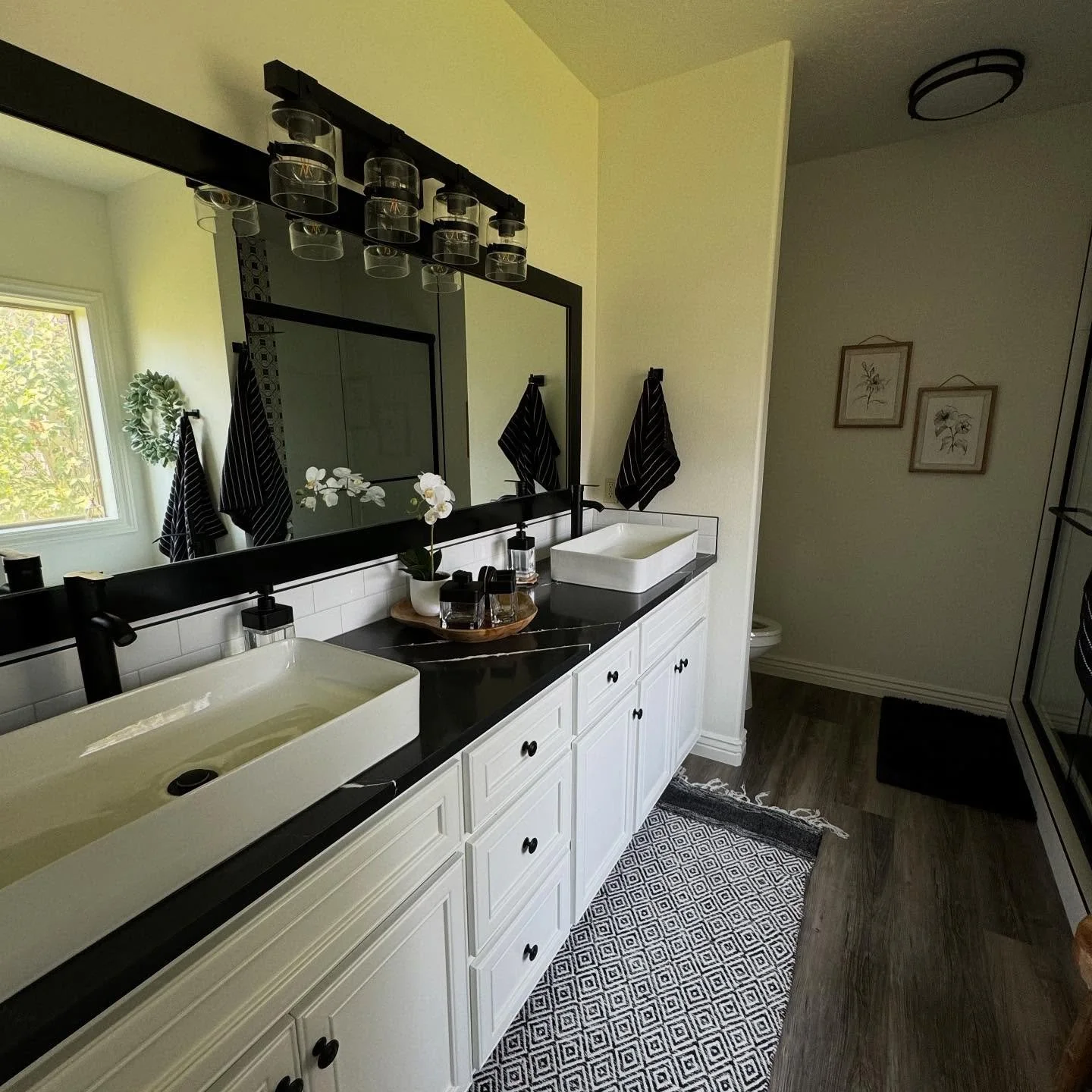 A bathroom with a large mirror, black countertops, and double white vessel sinks. There are two black towels hanging on hooks, a decorative tray with toiletries, a potted orchid, and a window letting in natural light. The floor has a patterned rug, and the bathroom has a light and neutral color scheme.
