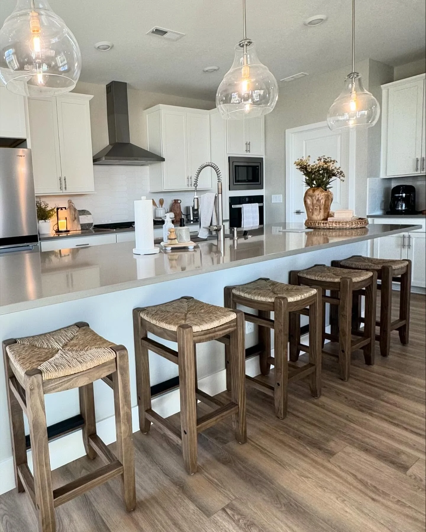 Kitchen with white cabinets, stainless steel refrigerator, black coffee maker, and decorated island with four wooden stools with woven seats.