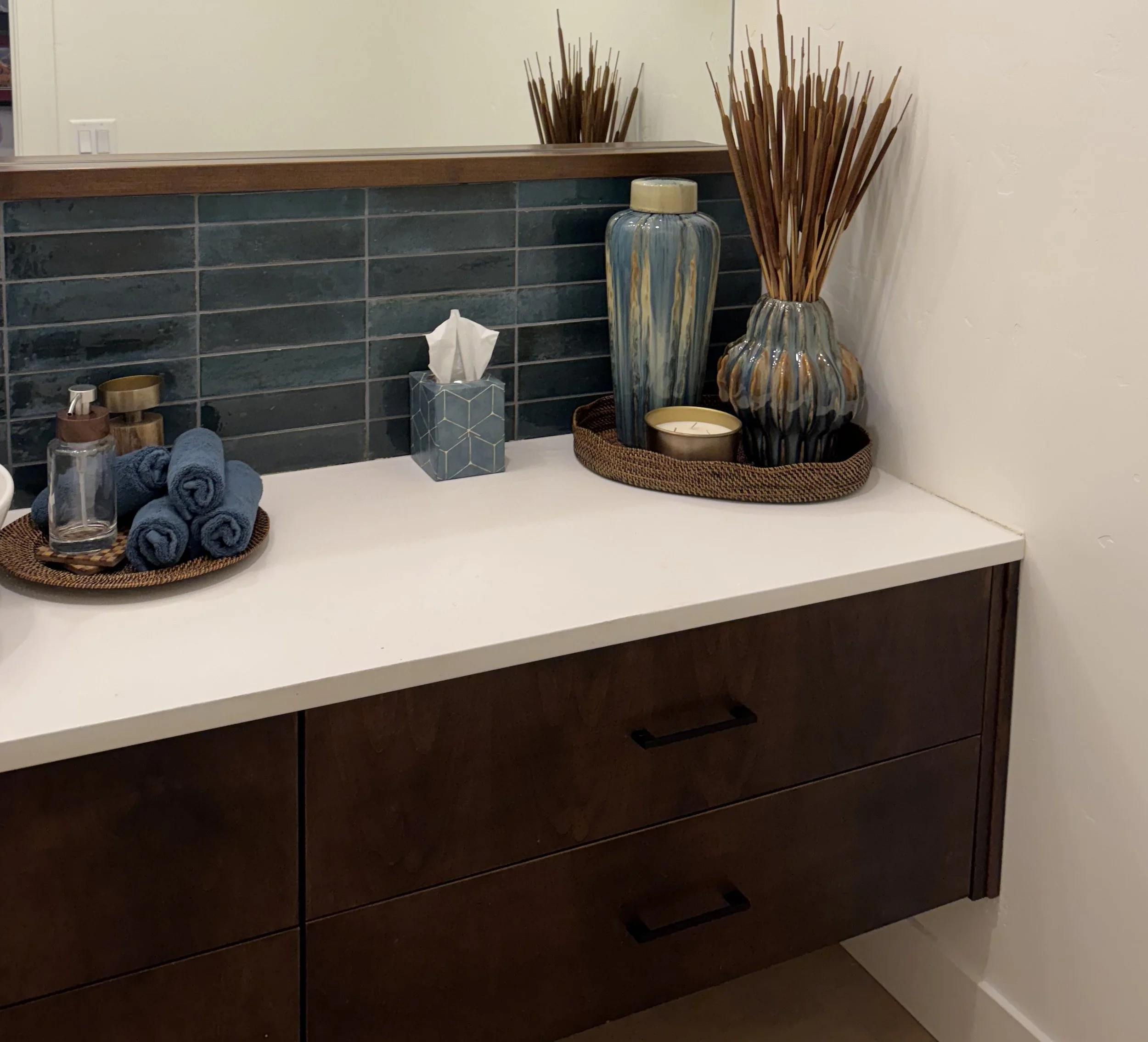 A bathroom vanity with a dark wood finish, a white countertop, and a large mirror. Decor includes tall vases with branches, a tissue box, a candle, and rolled towels.