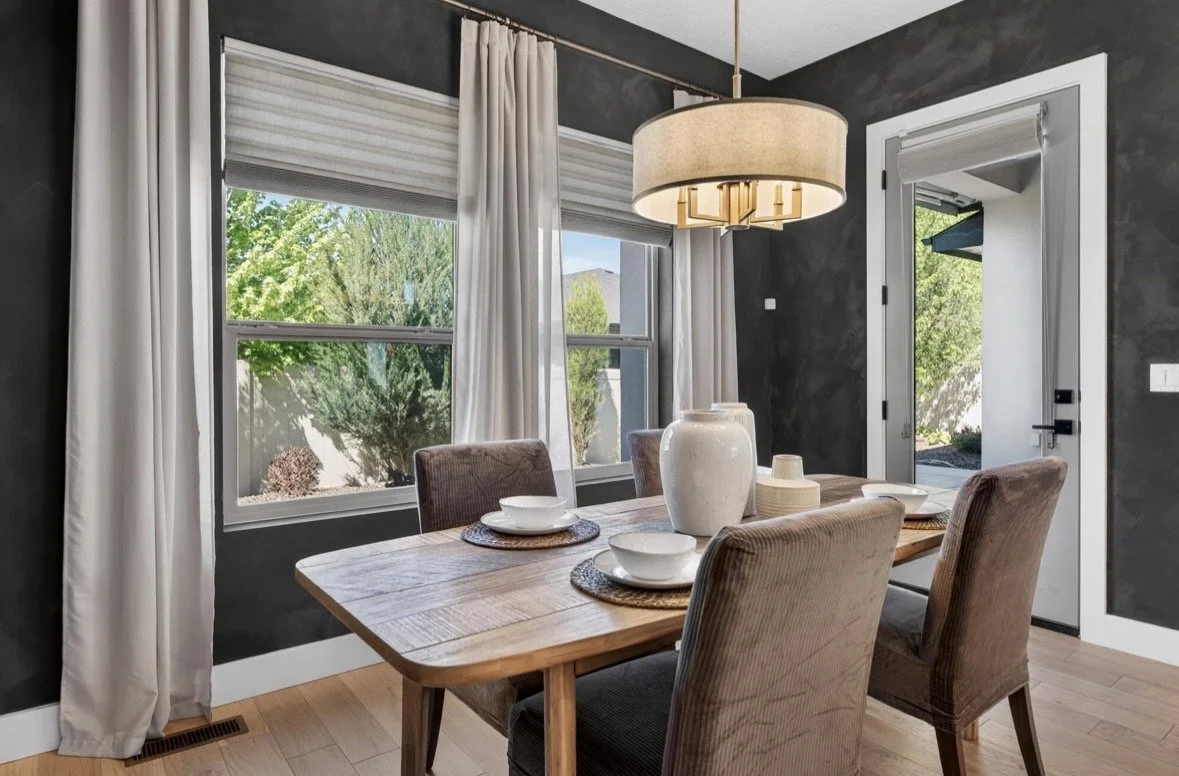 Dining room with a wooden table, four upholstered chairs, a large pendant light, and large windows with white curtains showing greenery outside.