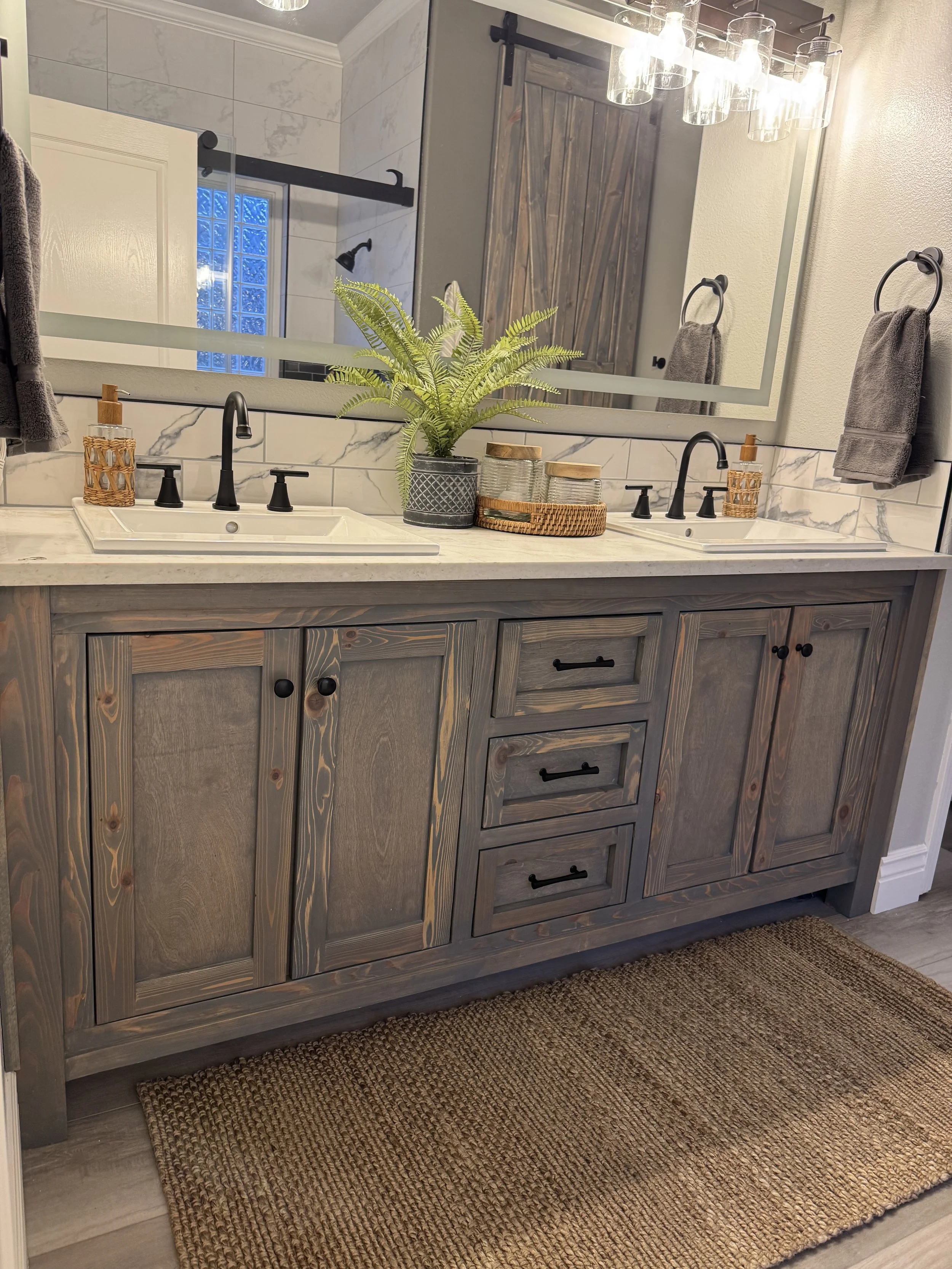A bathroom vanity with a double sink, gray wooden cabinets, black faucets, a large mirror, a potted fern, woven baskets, gray towels, and a textured brown rug.