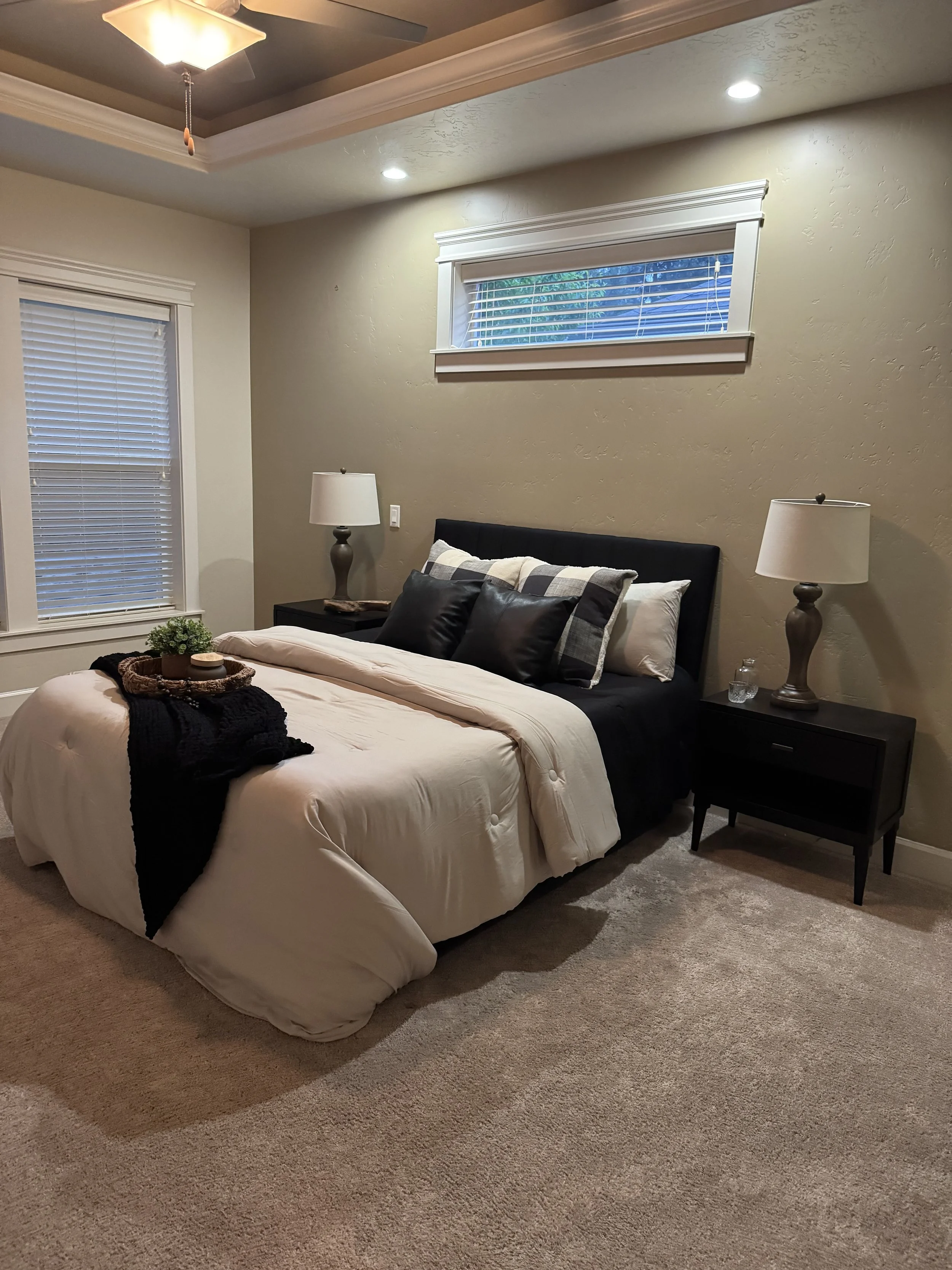 A bedroom with a bed, bedside tables with lamps, a window with blinds, and beige carpeted flooring.