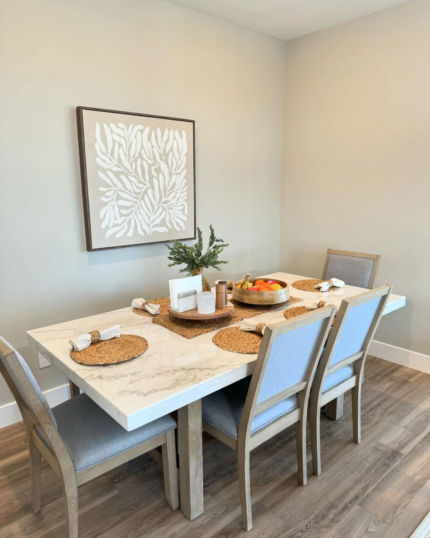 Dining room with a white marble table set with placemats, napkins, and a centerpiece with fruit. Six wooden chairs around the table, a wall with a framed white botanical art print, and a plant on the table.