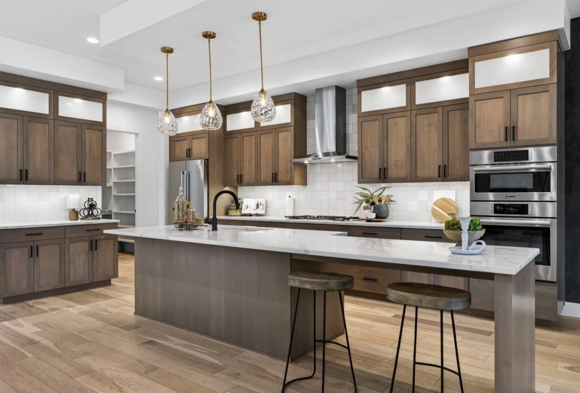 Modern kitchen with wooden cabinets, white countertops, and a large island with two stools. There are hanging pendant lights above the island, stainless steel appliances, and decorative items like plants and a tray with lemons.