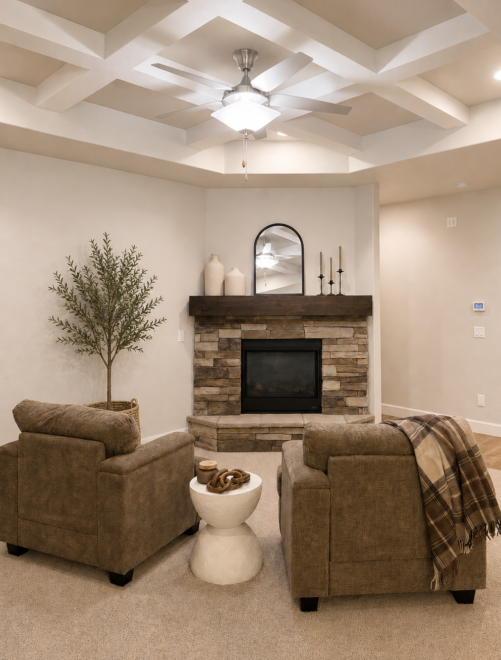 Living room featuring two upholstered armchairs, a stone fireplace with a dark wood mantel, a mirror, vases, candlesticks on the mantel, a small white side table with decorative objects, a potted plant, and a ceiling fan with a light in a coffered ceiling.