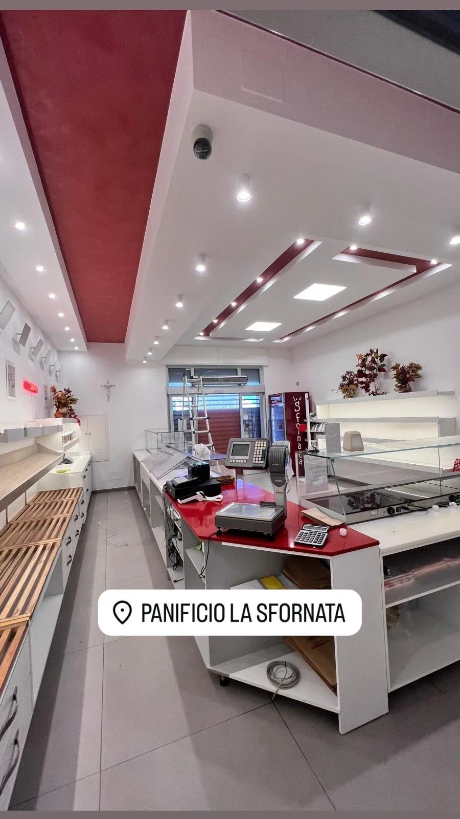 Interior of a bakery shop with a red and white counter, cash registers, and baking equipment. Shelves on the left and a ladder near the window. The ceiling has recessed lighting and decorative panels. Sign reads "Panificio La Sornata."