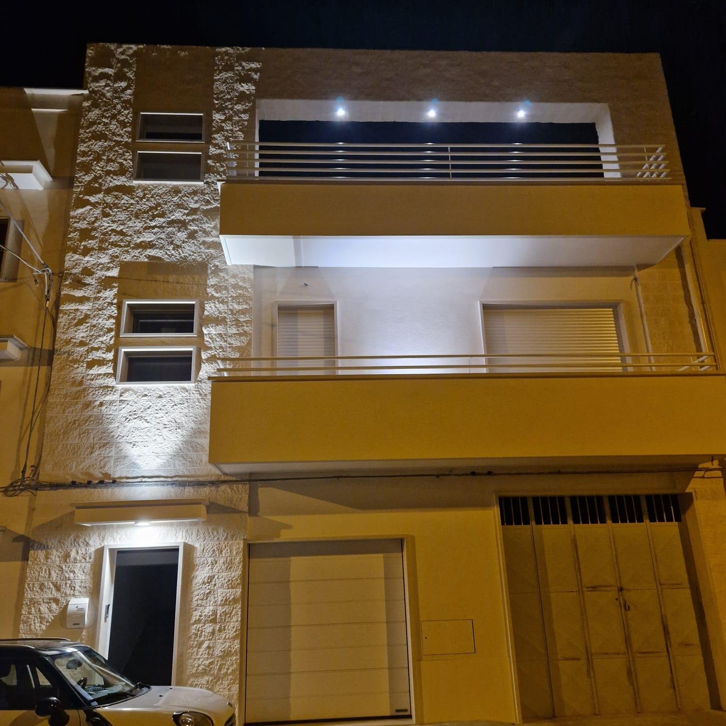 Nighttime view of a modern three-story residential building with yellow and stone exterior, multiple windows, balconies with railings, a garage, and a parked car in front.