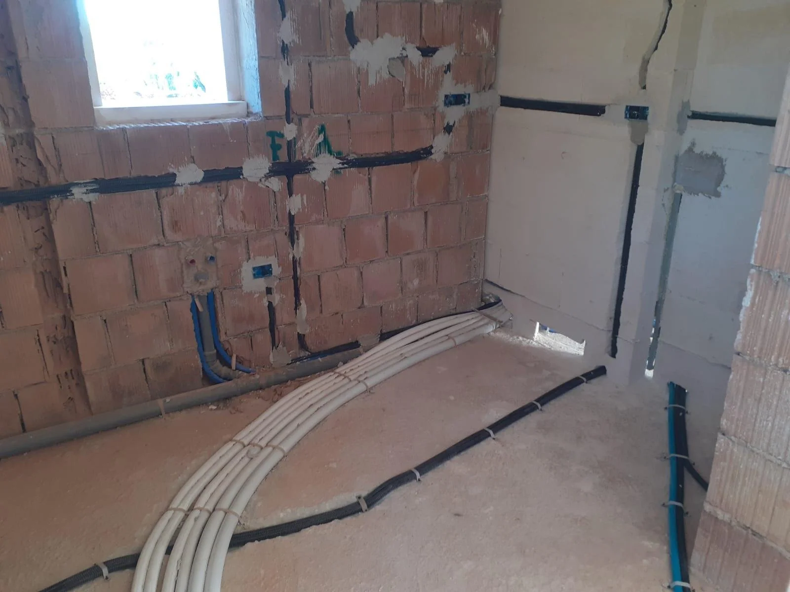 Interior construction site with exposed brick wall, electrical wiring and conduit pipes on the floor, and a window in the background.