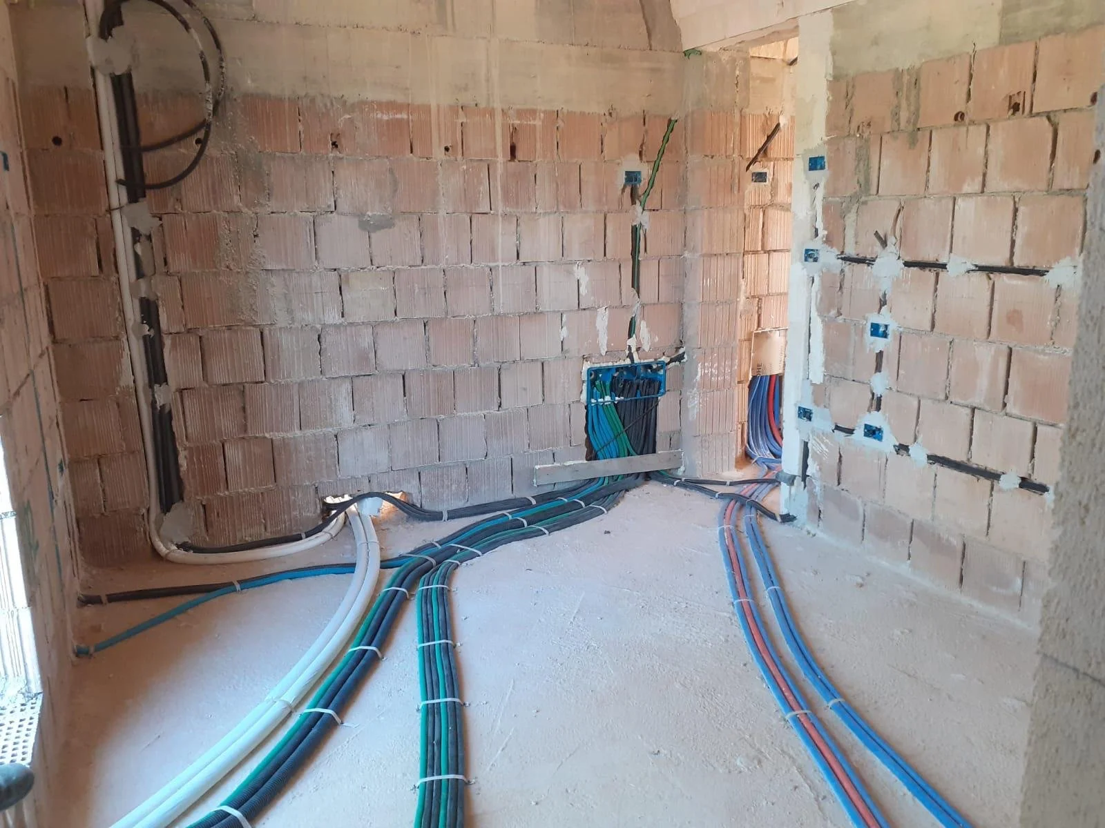 Interior view of a building under construction showing electrical wiring and conduit installation on exposed brick walls with various cables running across the floor.