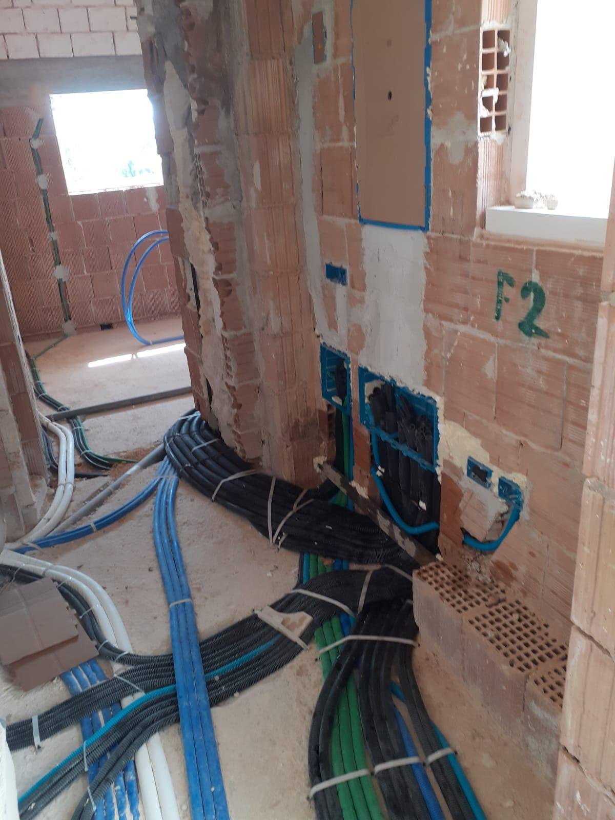 Inside a building under construction, exposed brick walls with electrical wiring, plumbing pipes, and conduits installed on the floor and wall.