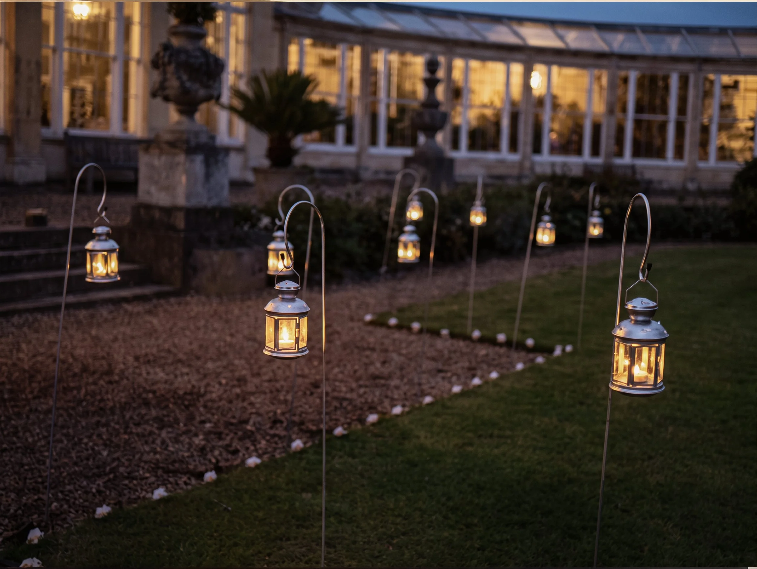 A garden at dusk with small hanging lanterns on curved hooks illuminating the walkway, a stone staircase with classical statues and planters, and a building with large windows glowing with warm light in the background.