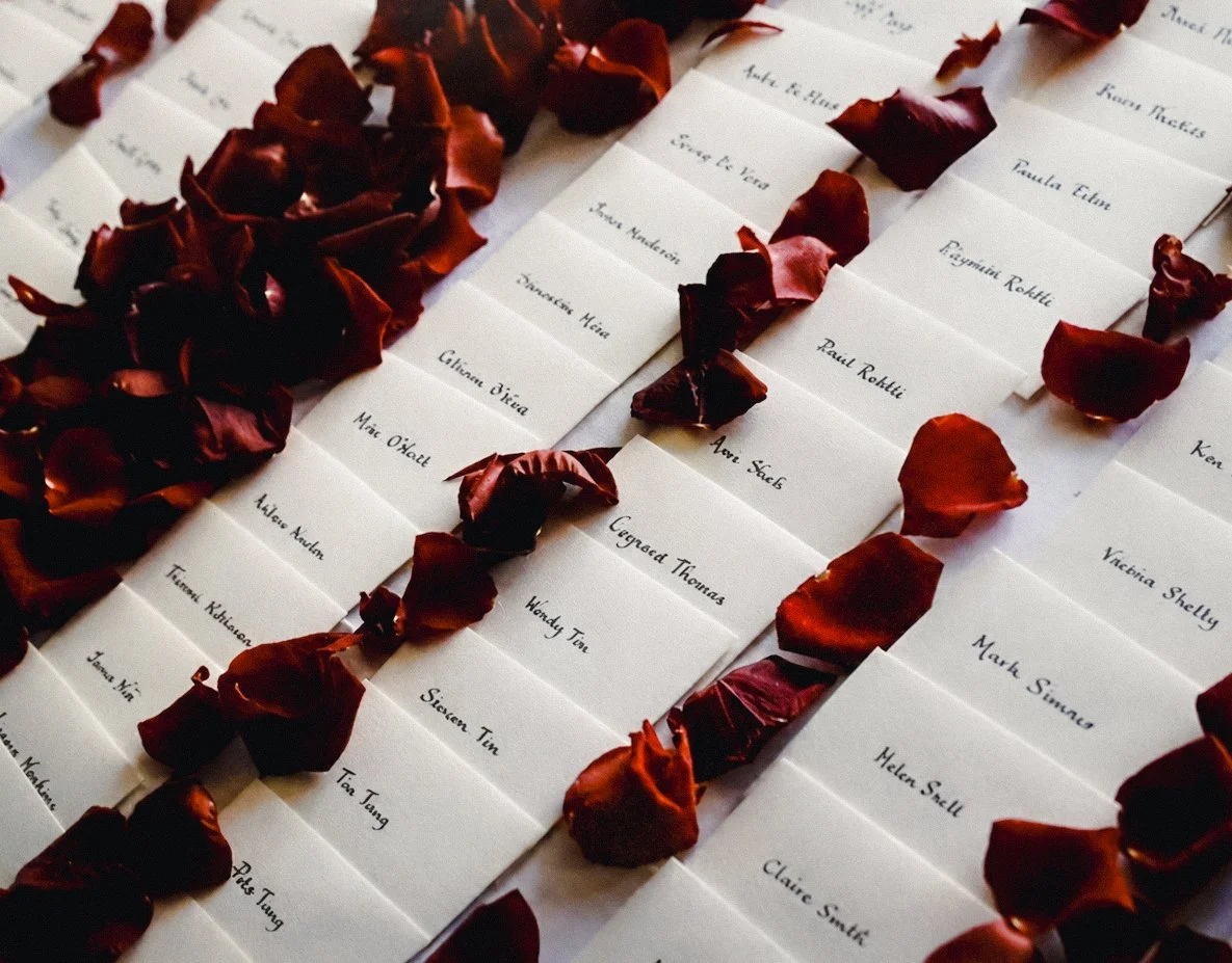 Names written on white cards laid out on a surface with dark red rose petals scattered around.