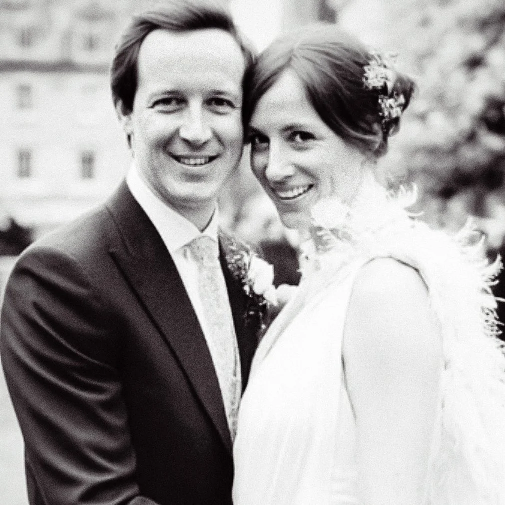A black and white photo of a happy couple, likely on their wedding day, posing outdoors. The man is wearing a suit and tie, and the woman is dressed in a wedding gown with hair adorned with a decorative hairpiece.