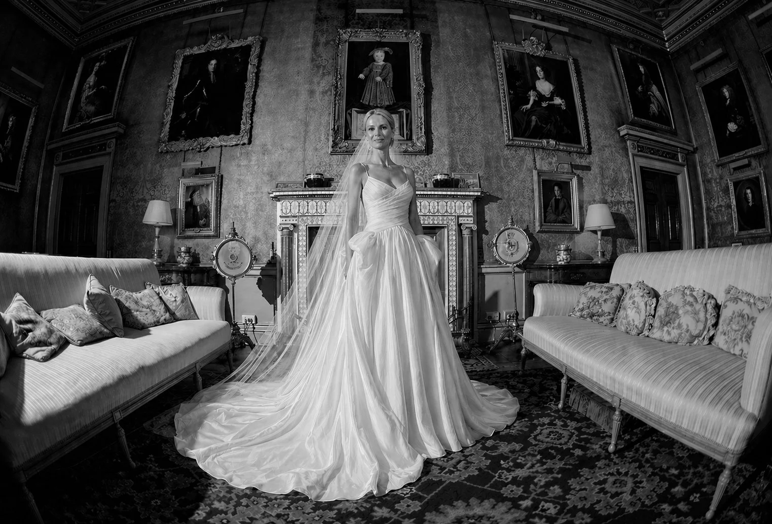 A woman in a wedding gown standing in an ornate, vintage living room with framed portraits on the wall and two striped sofas.