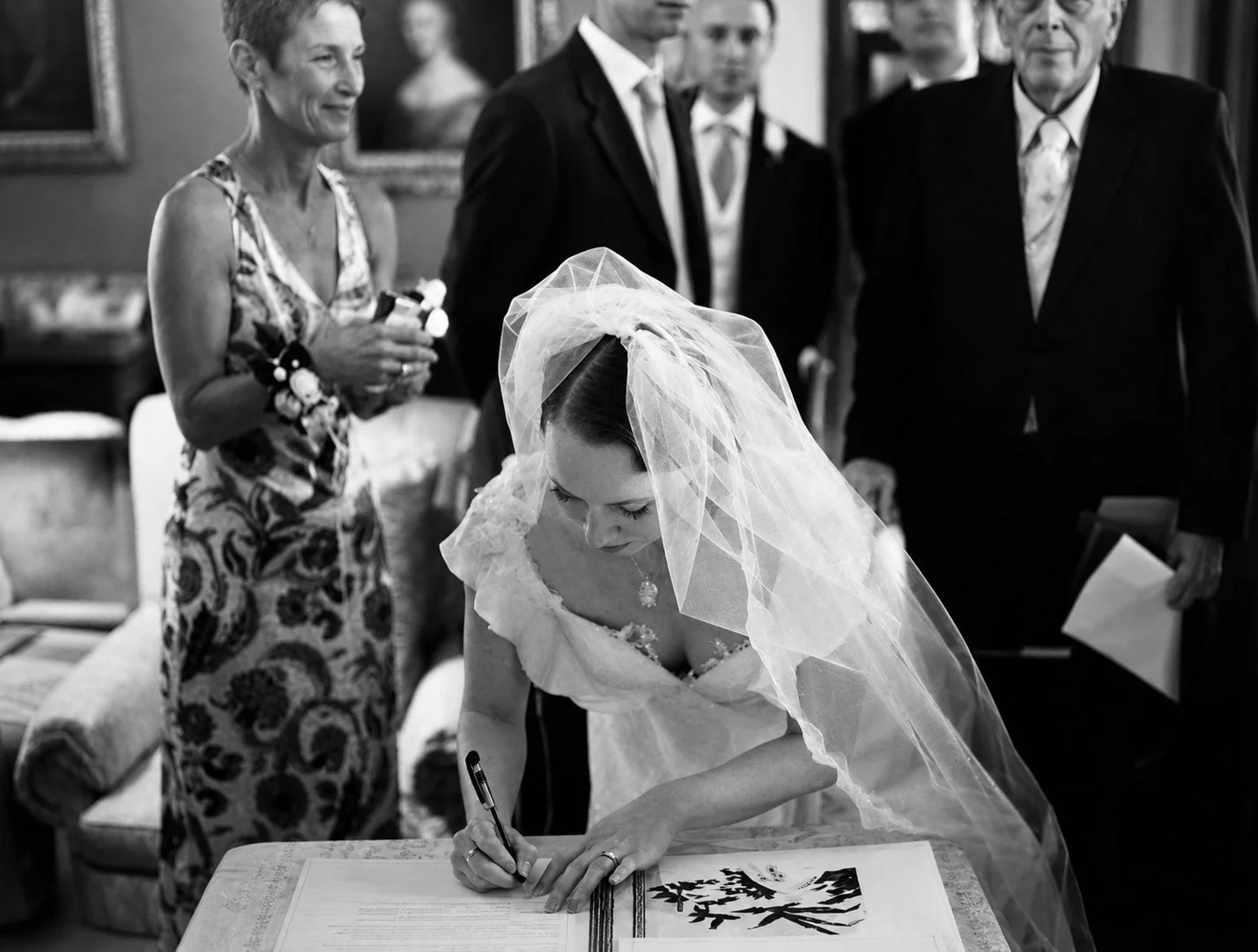 A bride wearing a wedding dress and veil is signing a document at a table, surrounded by four adults, including a woman in a patterned dress and three men in suits, in an indoor setting.