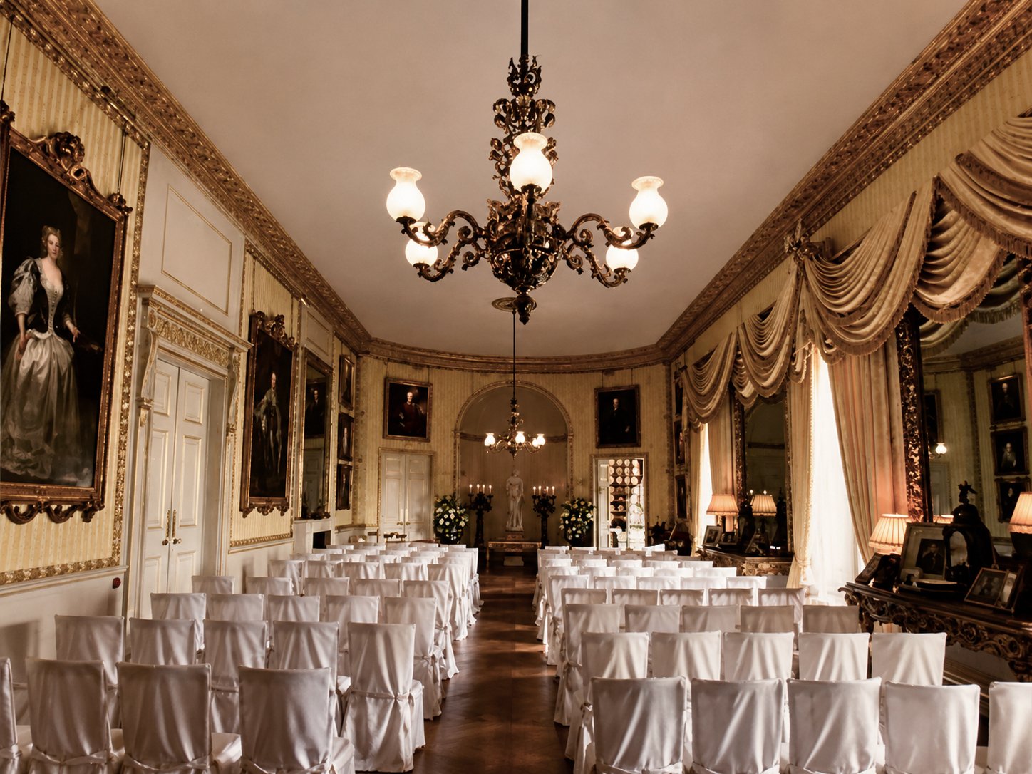 Elegant wedding or formal event room with ornate chandeliers, gold-trimmed artwork, and cream-colored draped curtains. Rows of white chairs face a stage with a statue and floral arrangements.