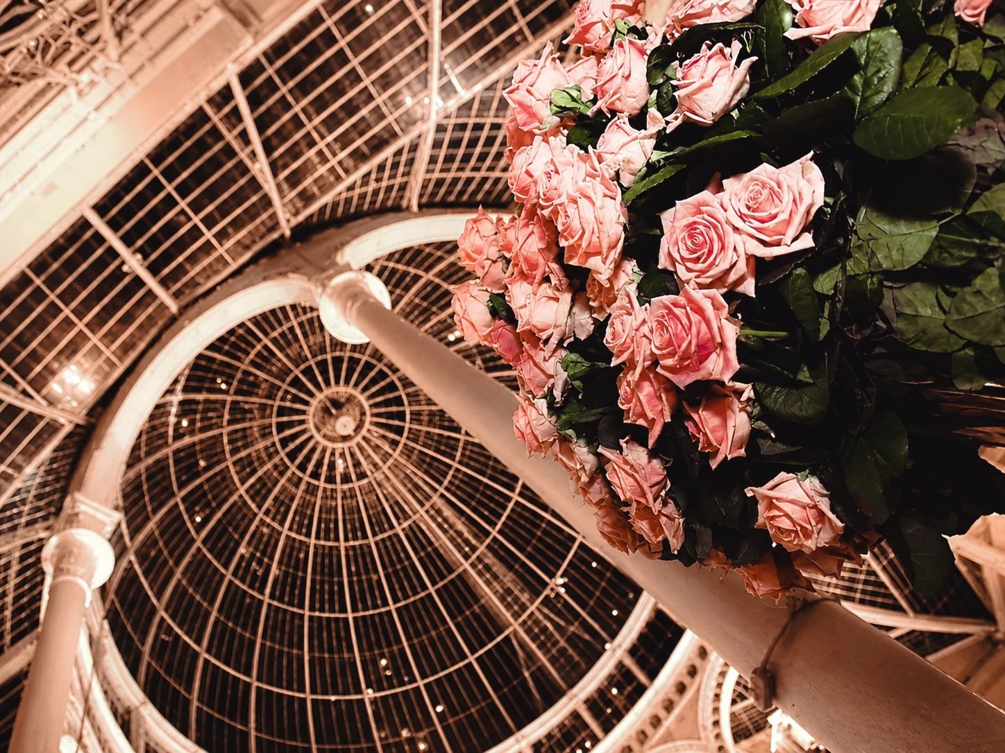 Looking up at a glass dome ceiling with a floral arrangement of pink roses and green leaves on a column in the foreground.