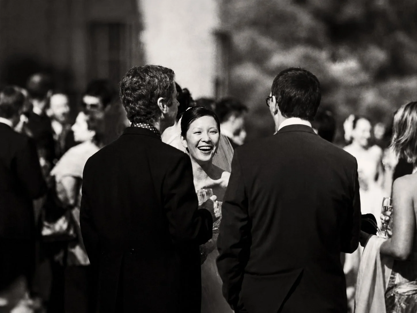 A black and white photo of a wedding reception with guests socializing, with a woman in a wedding dress smiling and talking to two men in suits.