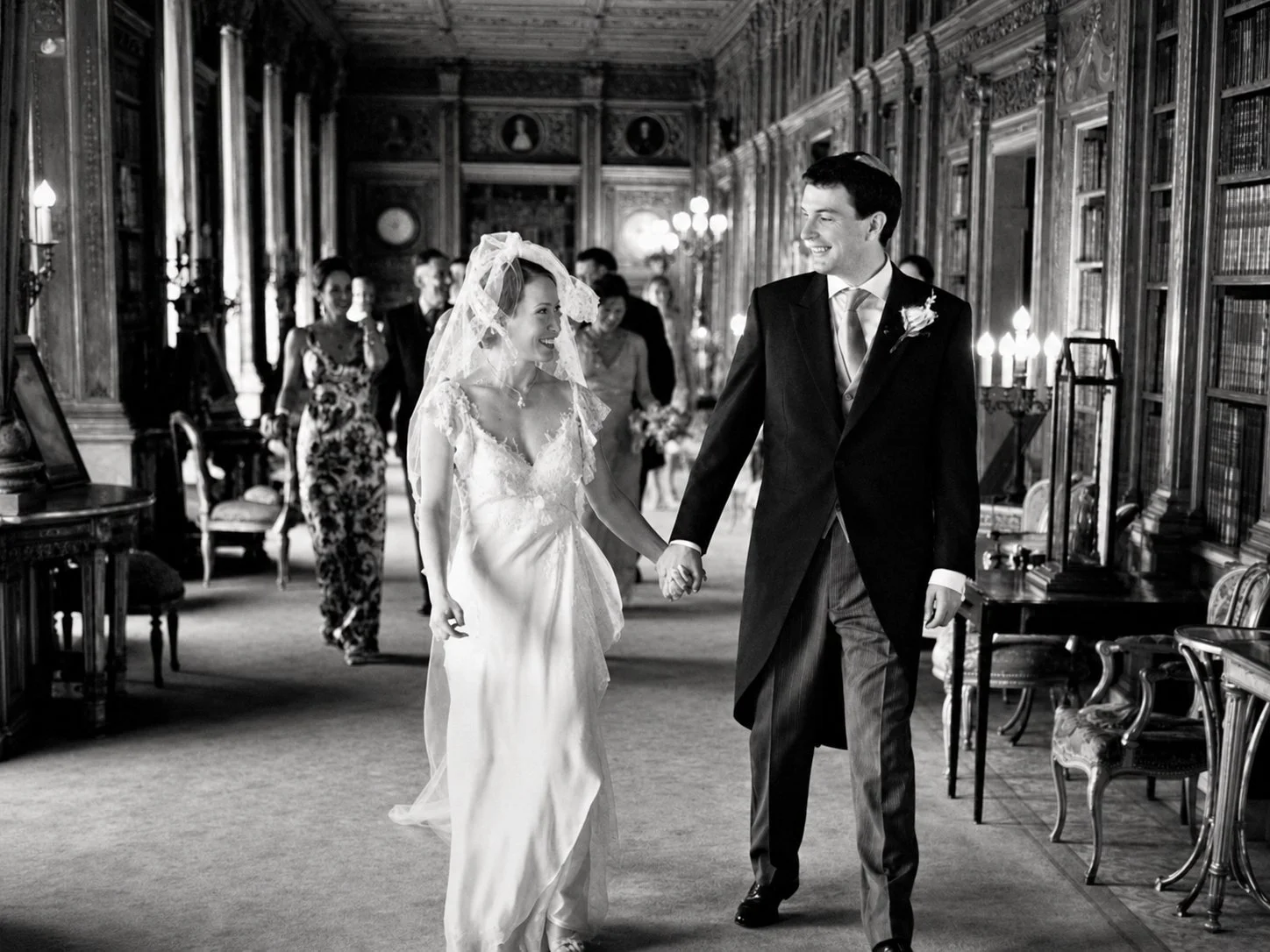A bride and groom holding hands and smiling at each other in a historic wood-paneled room during their wedding. The bride is wearing a wedding gown with lace details and a veil, and the groom is dressed in a suit with a boutonniere. Several guests ar
