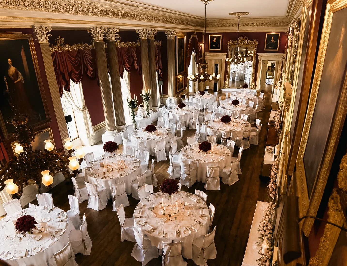 Elegant banquet hall with round tables covered in white tablecloths, each decorated with dark red floral centerpieces, set for a formal event, featuring tall curtains, grand columns, framed portraits, and ornate gold and white decor.