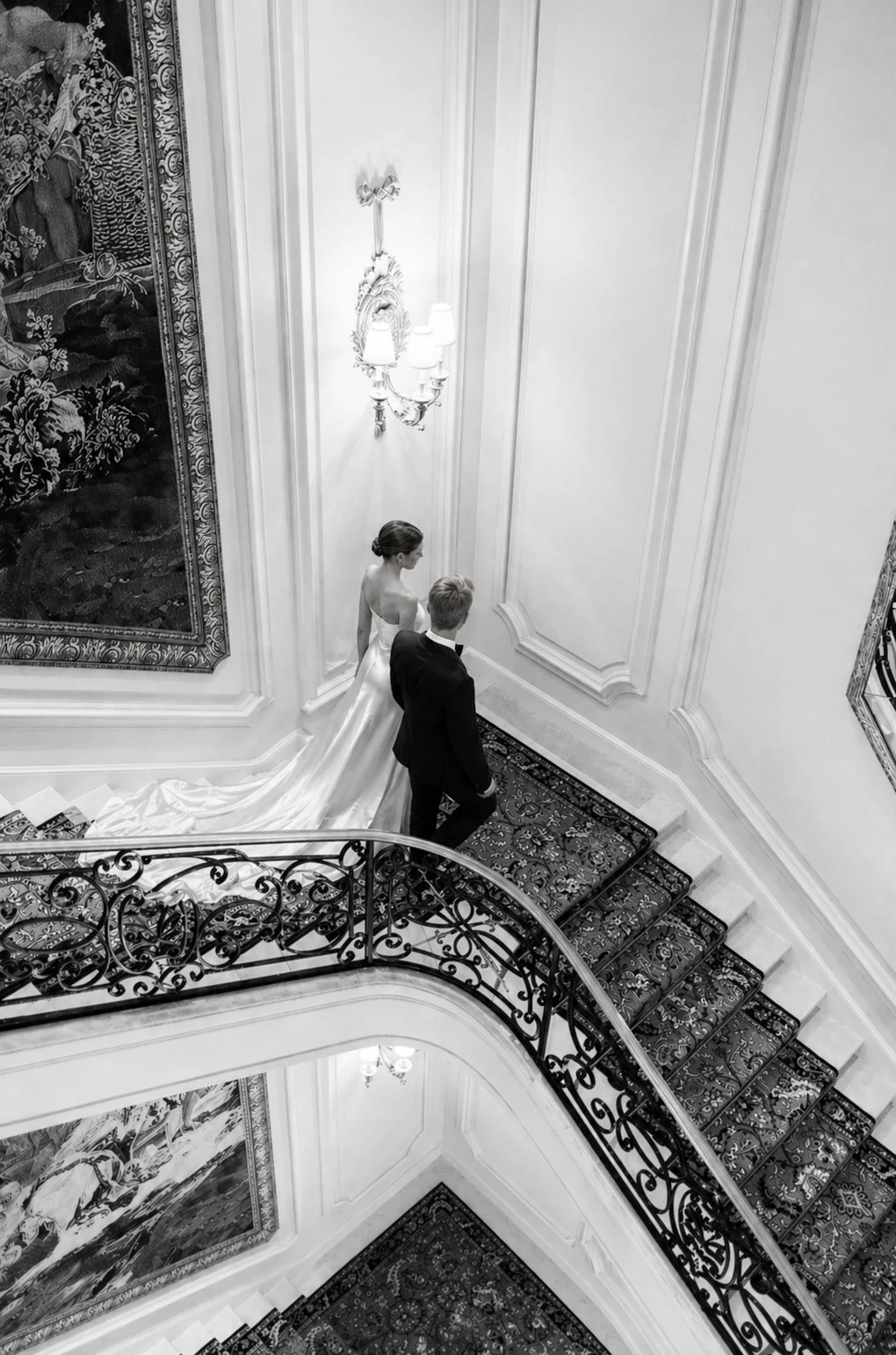 Black and white photo of a bride and groom walking down an elegant staircase in a grand, ornate interior. The bride is wearing a strapless wedding gown with a long train, and the groom is in a black suit. The staircase has intricate iron railings, an
