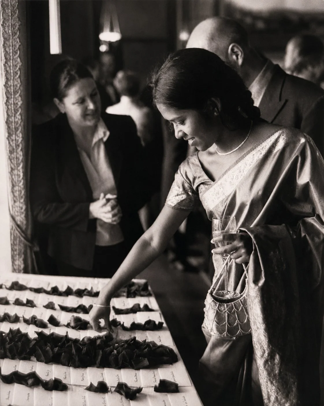 A woman in traditional attire reaching for chocolates or small items on a display table at a social event, with a man and another woman observing nearby, in a black and white photo.