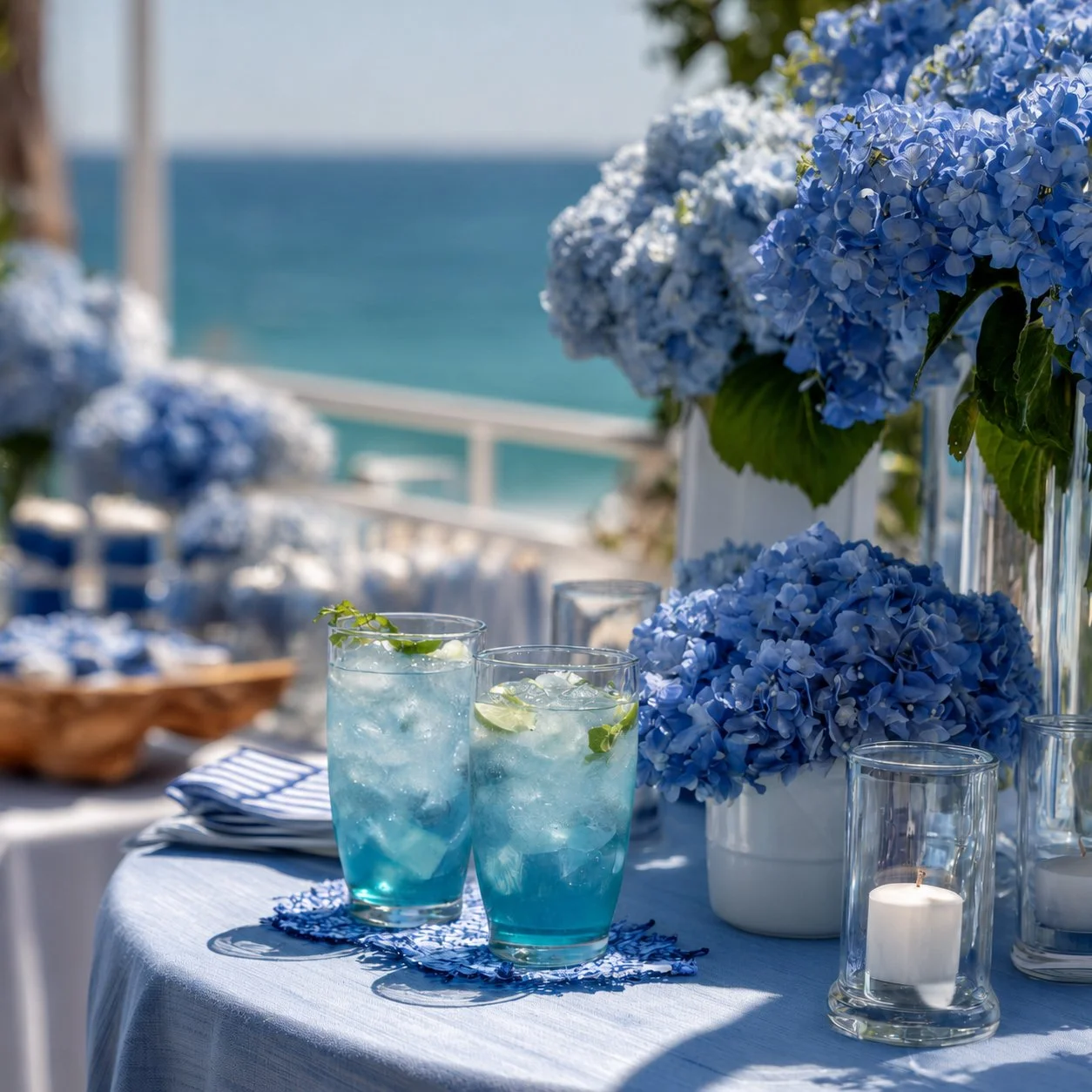 A table decorated with blue hydrangea flowers, two blue drinks with ice and lime slices, candles, and blue and white striped napkins, overlooking the ocean.