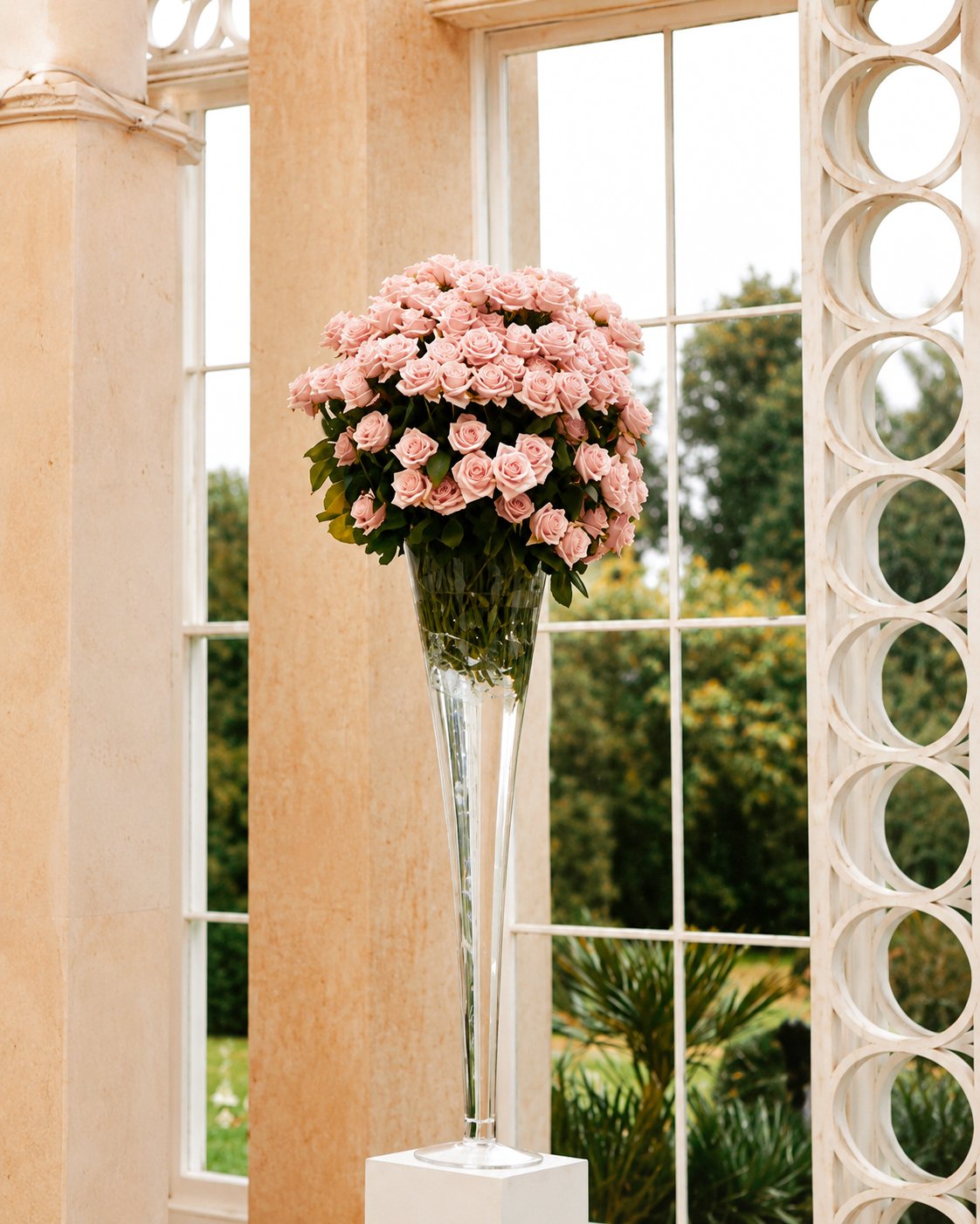 A large bouquet of pink roses in a tall, clear glass vase inside a room with large windows and decorative white architectural elements, with a view of greenery outside.
