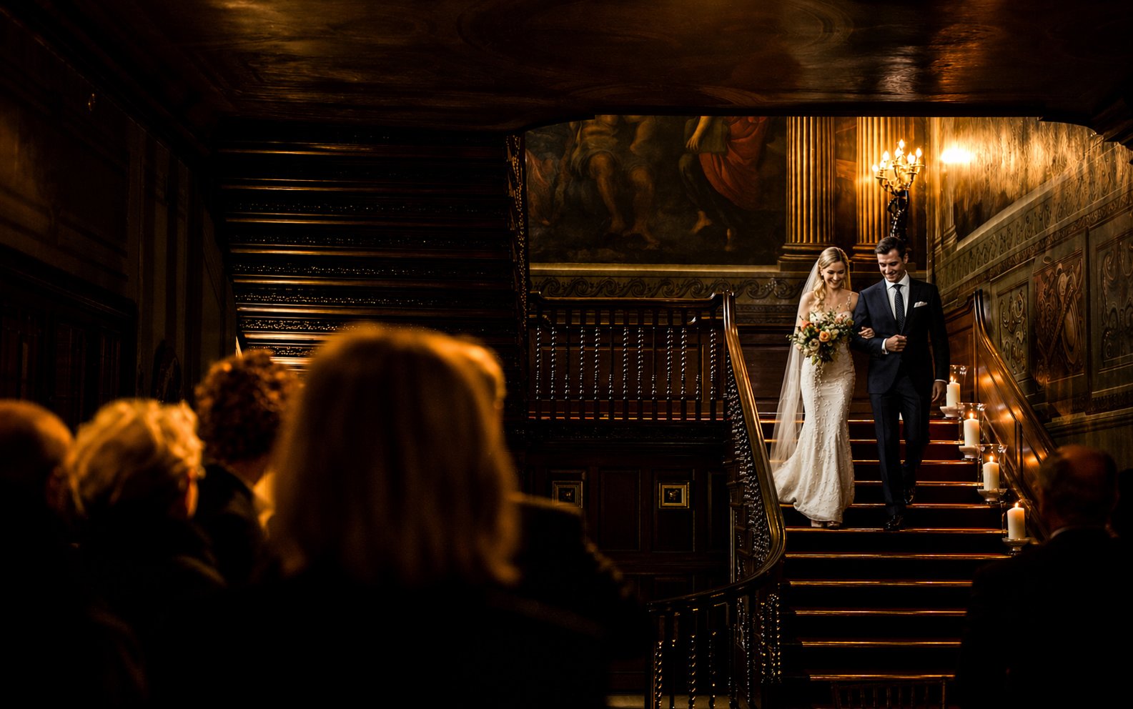 A bride and groom walking down a staircase during their wedding ceremony, with guests seated watching. The setting is an elegant, dimly lit interior with candles and ornate decorations.