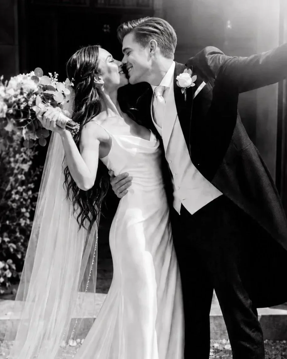 Black and white photo of a bride and groom sharing a kiss. The bride holds a bouquet of flowers, and the groom's arm is extended in a celebratory gesture. They are dressed in formal wedding attire.