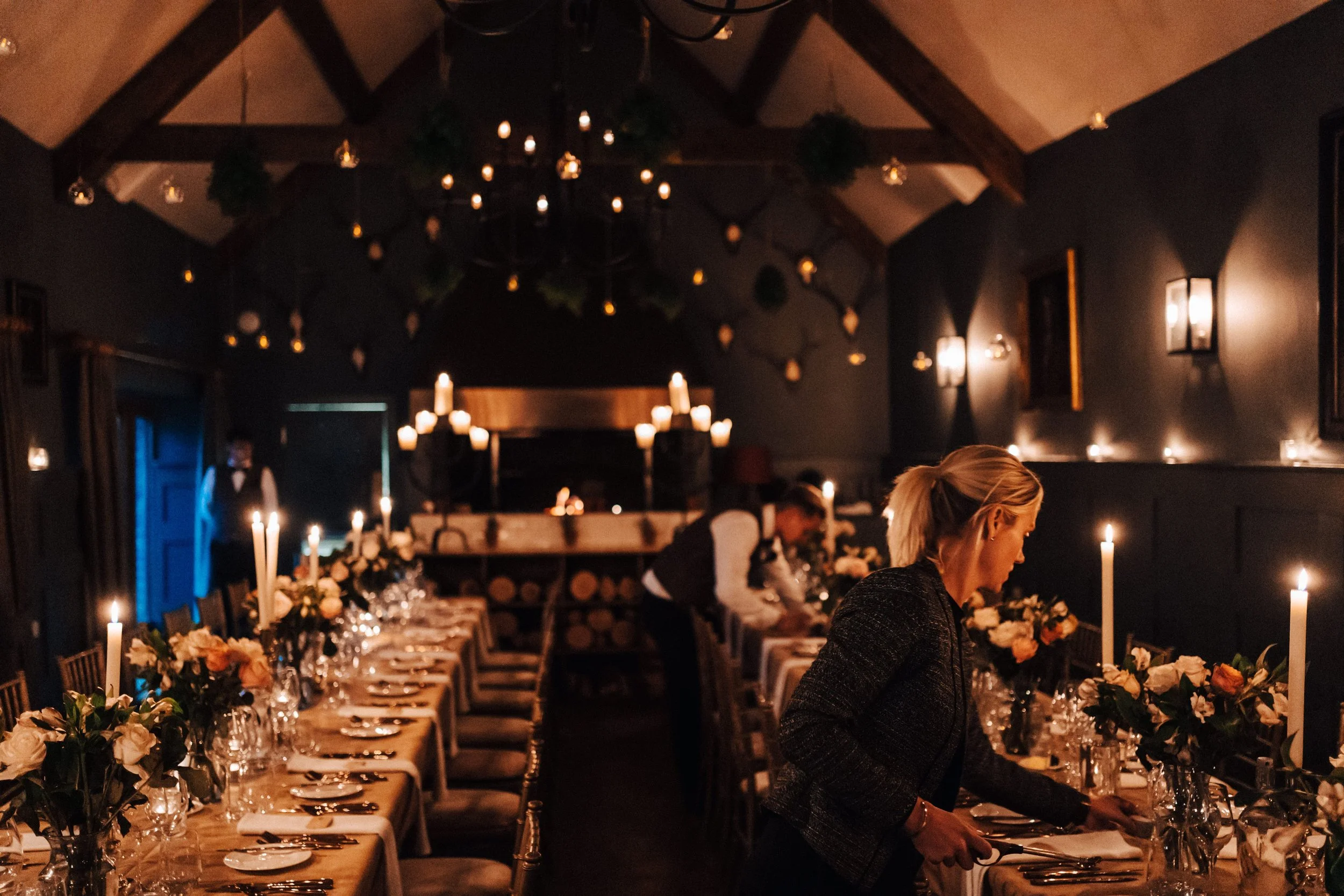 A dimly lit banquet room with long tables set with elegant floral centerpieces, candles, and fine dinnerware, decorated for a formal event, with waitstaff preparing the table.