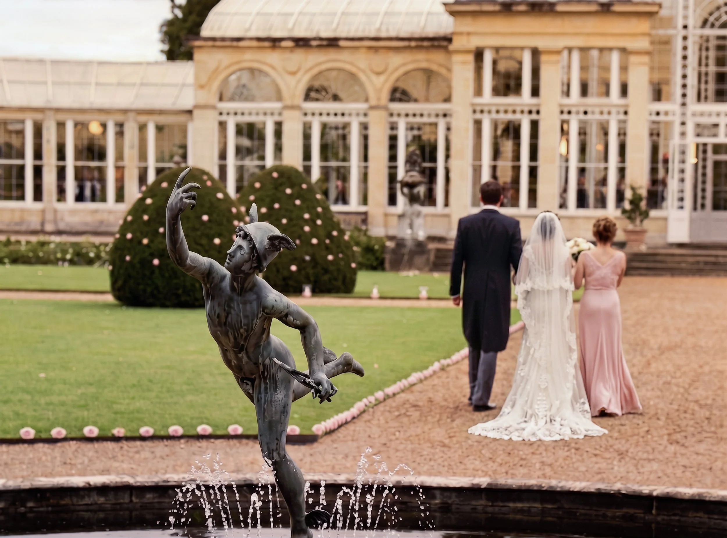 A statue of a young woman dancing is in the foreground of a wedding scene at a garden with a building in the background. The bride and groom are walking away from the camera, holding hands. The bride is wearing a white wedding dress and veil, and the