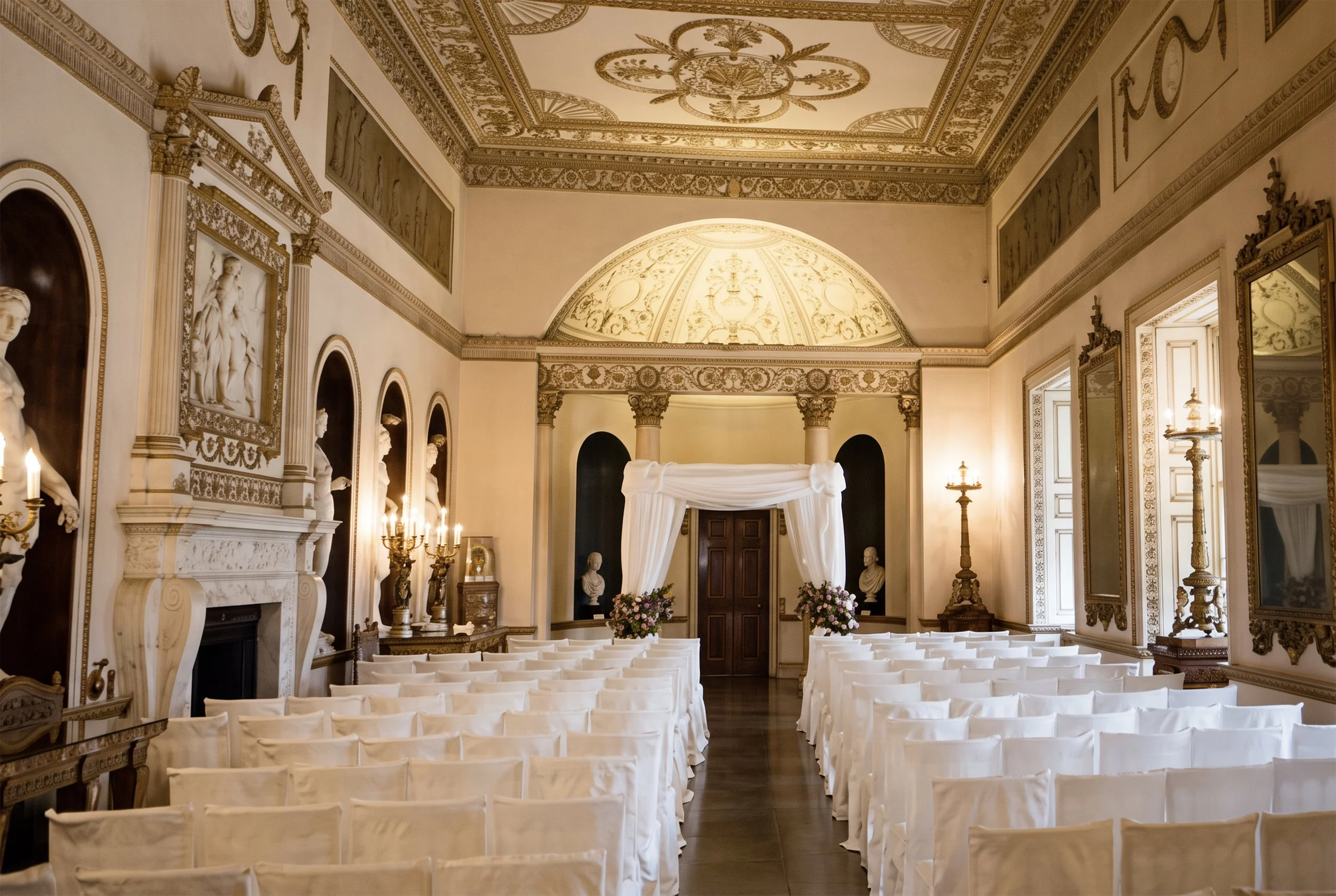 Artisan by EDLO luxury wedding design Elegant, historic wedding venue at Syon Park with ornate gold detailing on ceiling and walls, white draped arch at altar, and rows of white chairs awaiting a ceremony