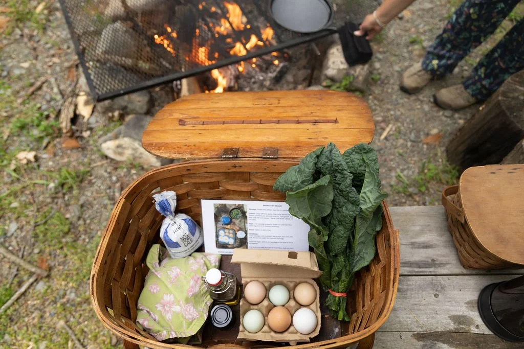 Basket containing fresh eggs, leafy greens, a bottle of oil, and other items, set on a wooden table outdoors near a campfire.