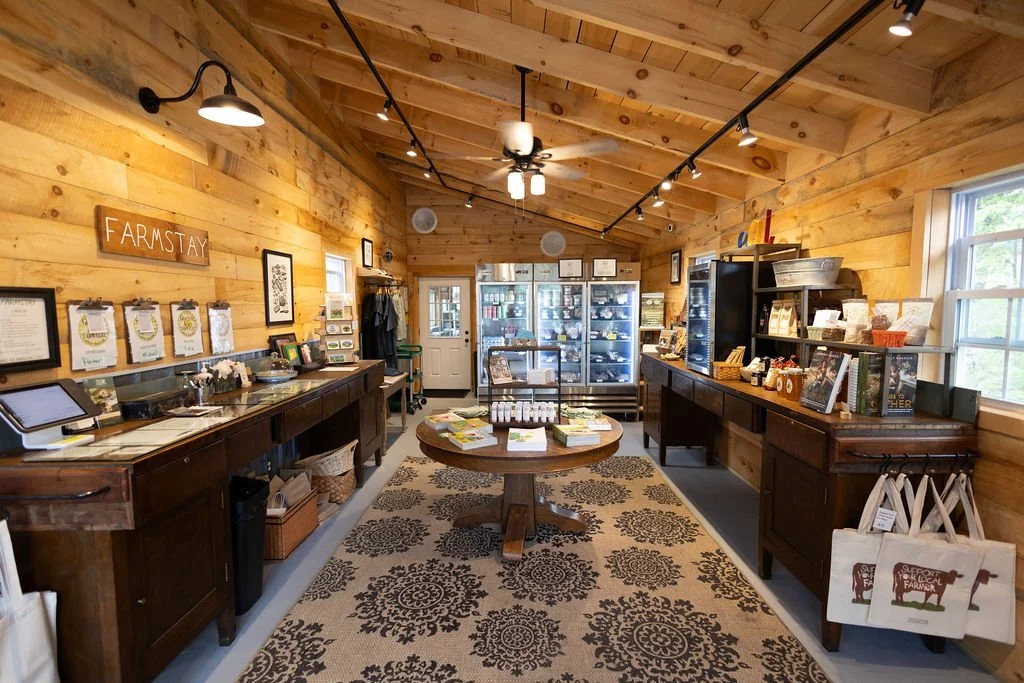 Interior of a rustic farm store with wooden walls and ceiling, displaying shelves of items, a central display table, and a patterned rug.