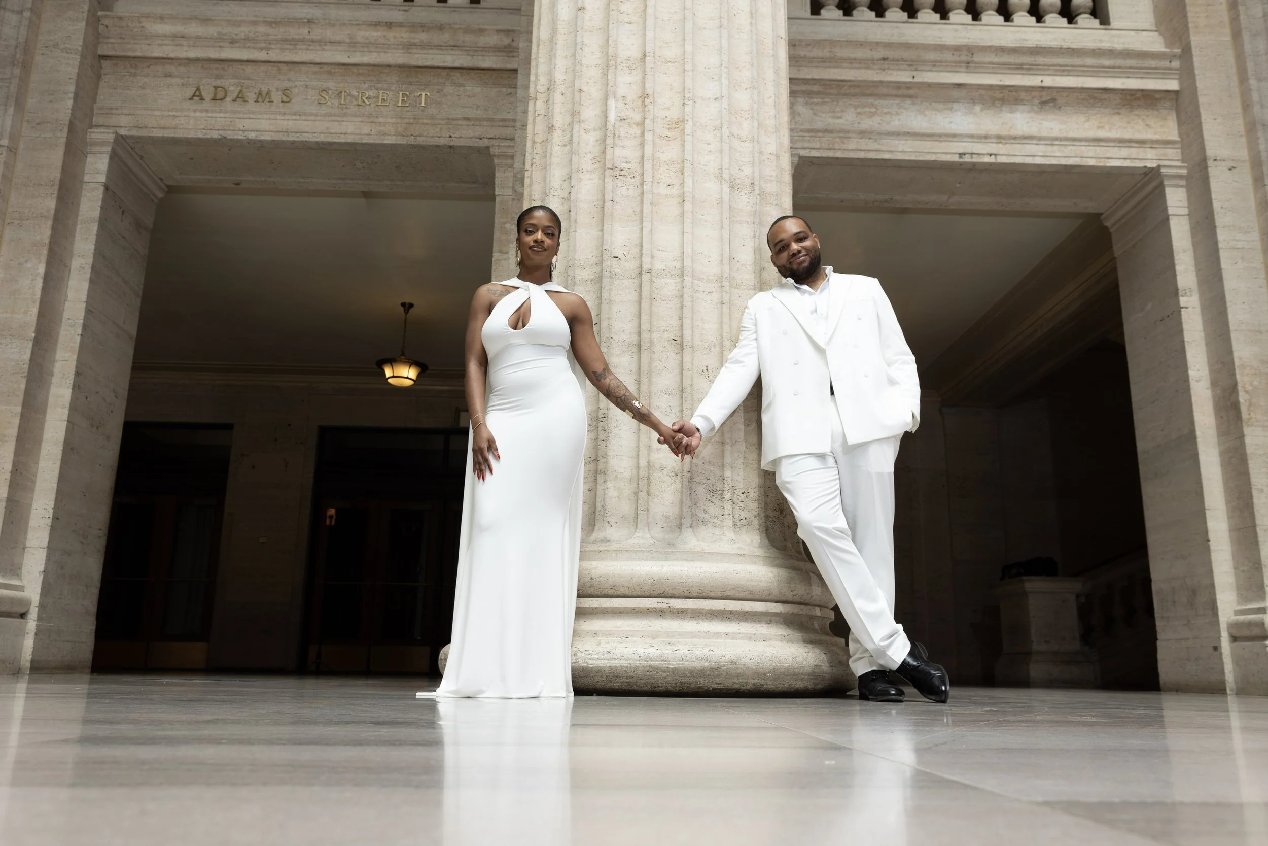 A couple holding hands standing in front of a stone building entrance with columns, with 'Adams Street' engraved above them, both dressed in white formal attire, smiling.