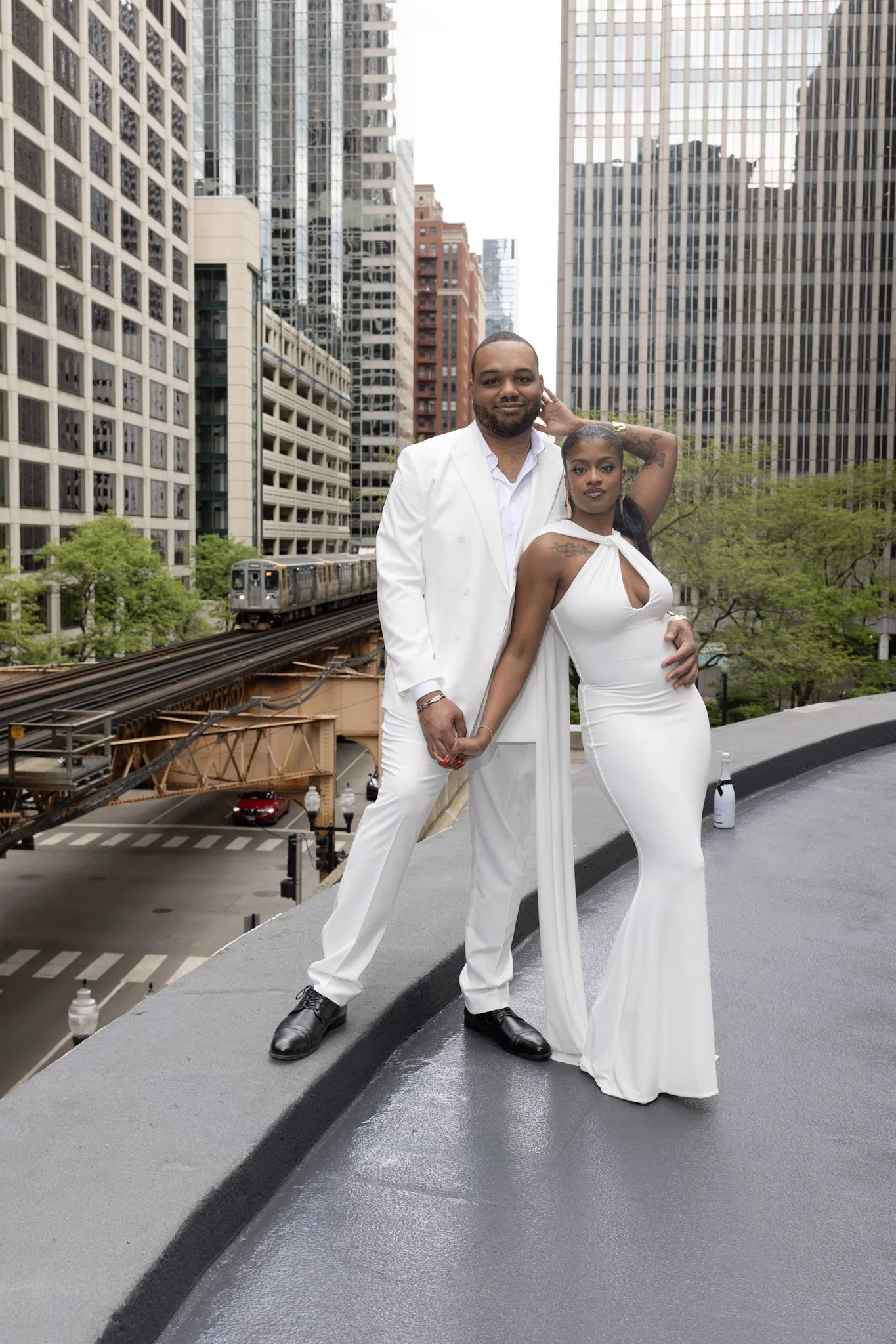 A man and a woman in white clothing standing on a city rooftop with tall buildings and a train in the background.