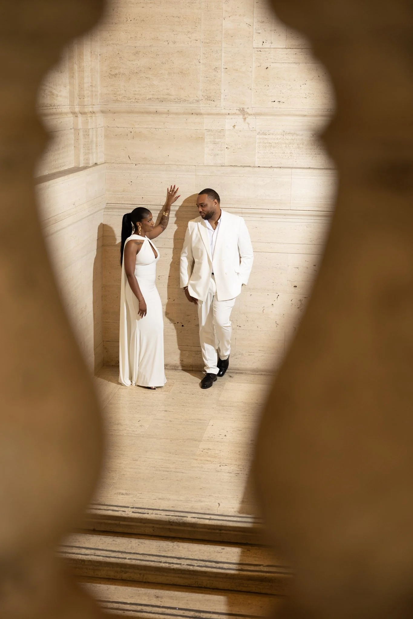 A man and woman dressed in white, standing against a beige marble wall, viewed through an ornate wooden frame.