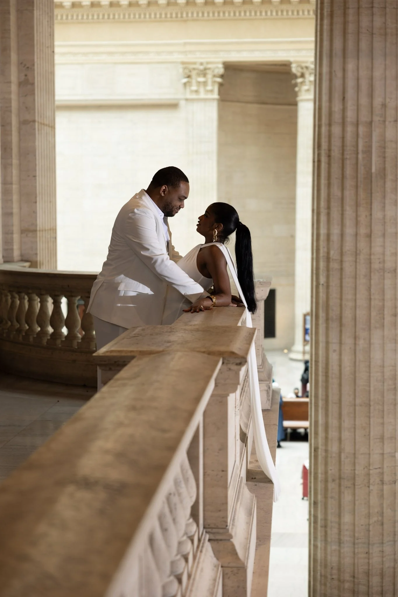 A couple dressed in white, standing close together on a balcony inside a grand building with classical architecture, including tall columns and decorative details, with a view of the foyer or interior space below.