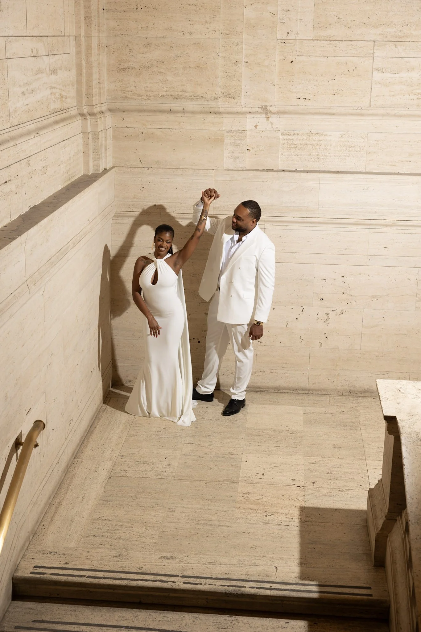 A man and woman in formal white attire celebrating, standing against a beige stone wall, with the woman raising her hand and the man holding her wrist.