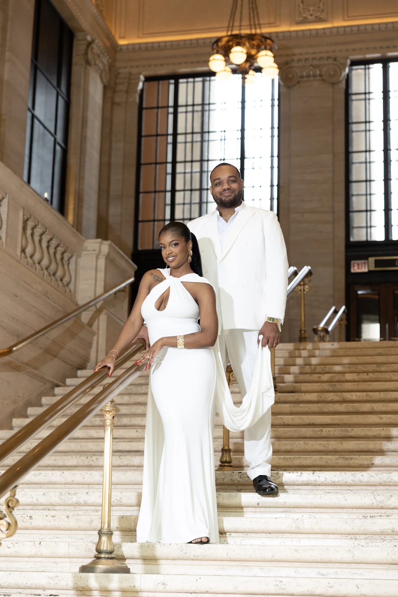 A stylish couple dressed in white attire standing on the grand staircase of an elegant building with high ceilings, large windows, and ornate details. The woman is leaning on the handrail with a confident smile, while the man is standing behind her with a slight smile, wearing a white suit.