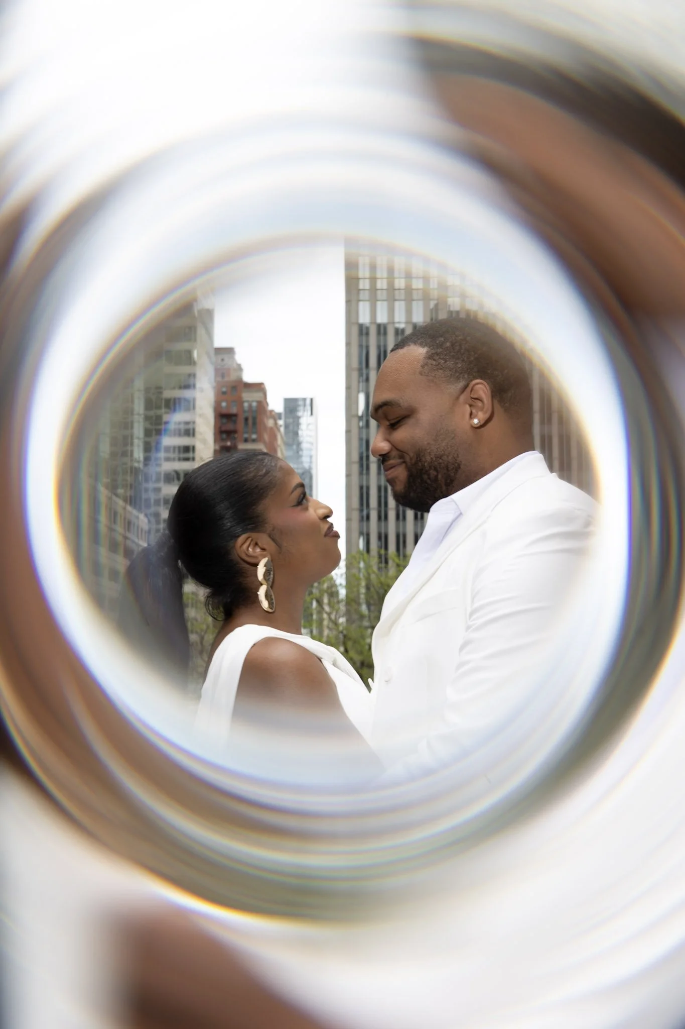 A man and woman facing each other, smiling in an urban cityscape with tall buildings, viewed through a circular frame or tunnel.